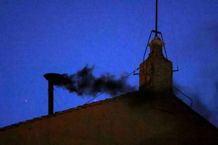 A chimney emits black smoke against a darkening sky, indicating a significant event, such as a papal election, in progress