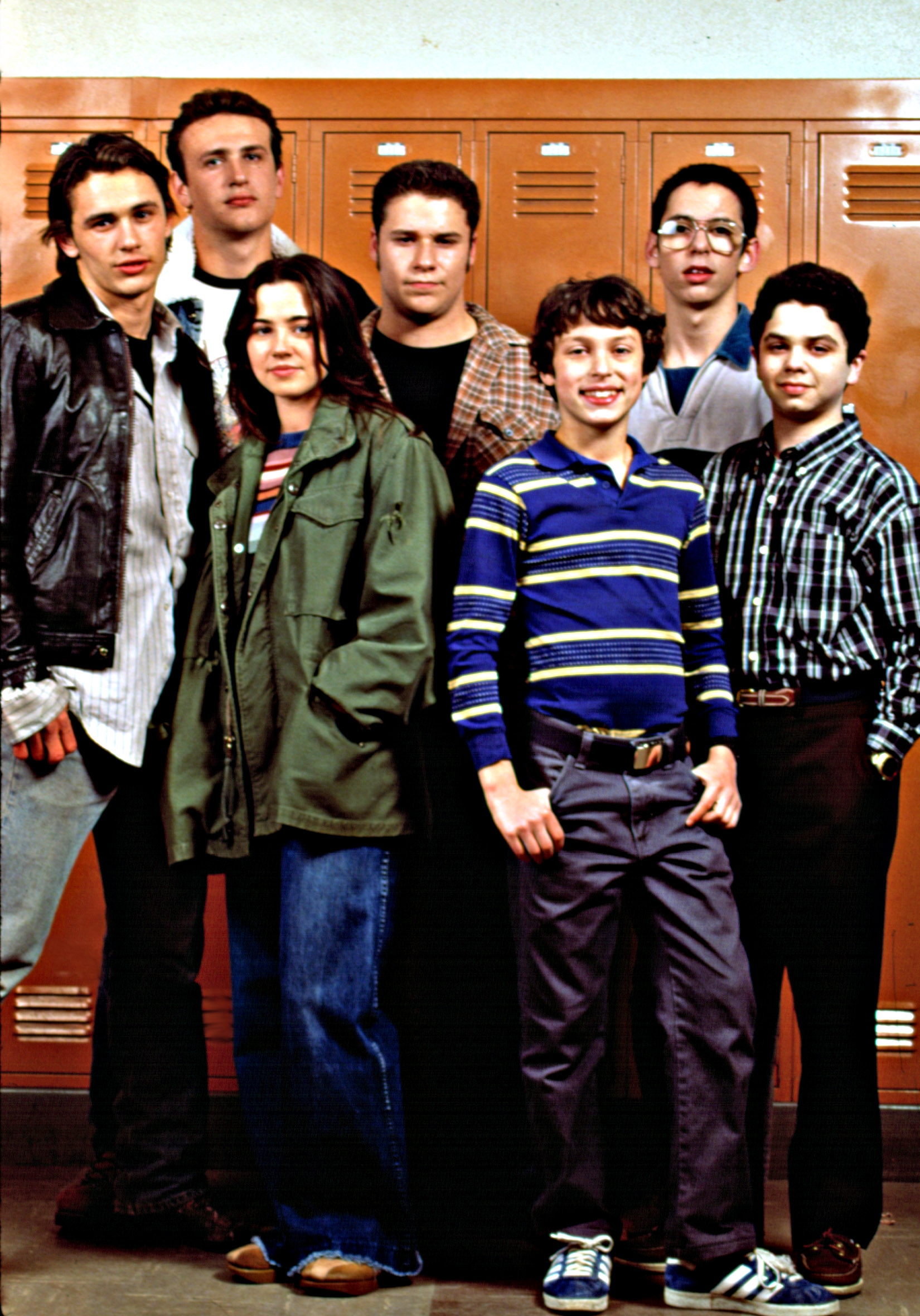 A group of young adults stands in front of lockers, wearing casual late 1990s clothing, including jackets and patterned shirts