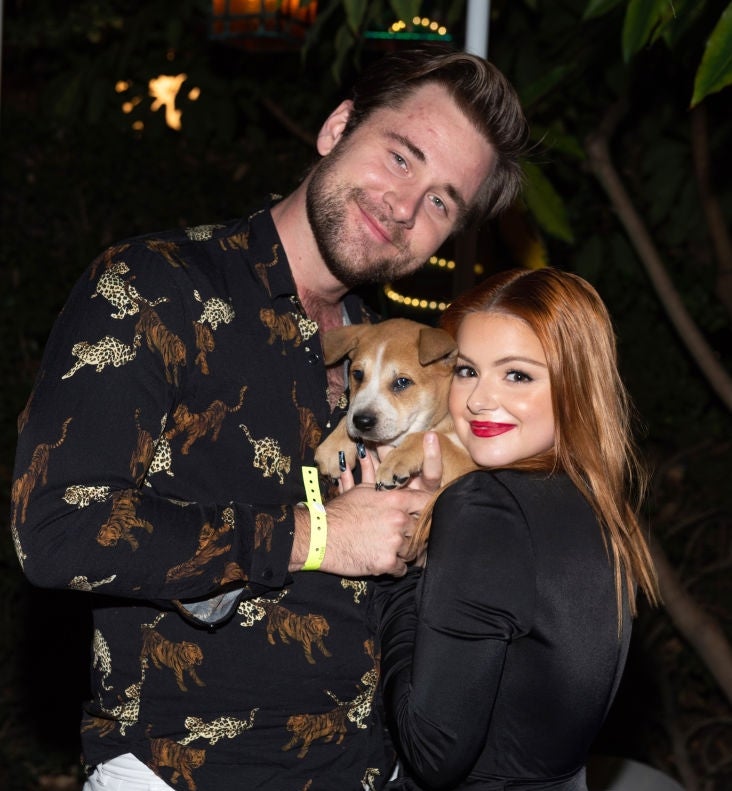 Two people smiling, holding a small dog. One wears a patterned shirt and white pants; the other wears a sleek, black dress