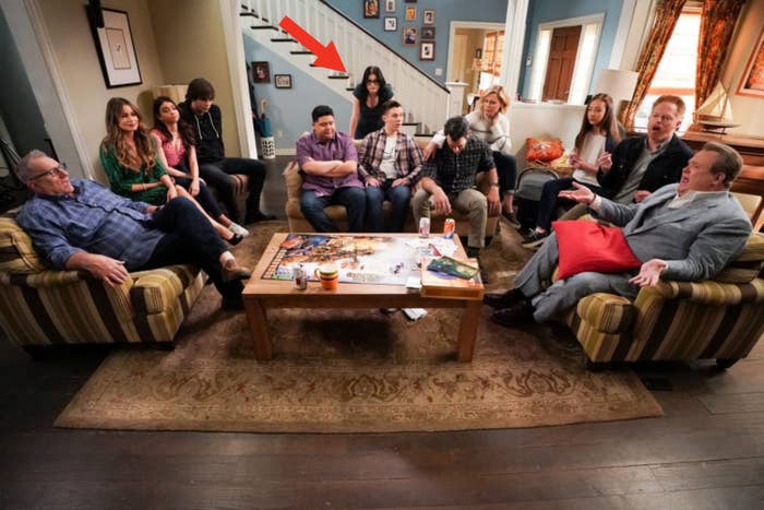 A group of people sit on couches and chairs in a living room, talking and interacting around a coffee table with board games and snacks