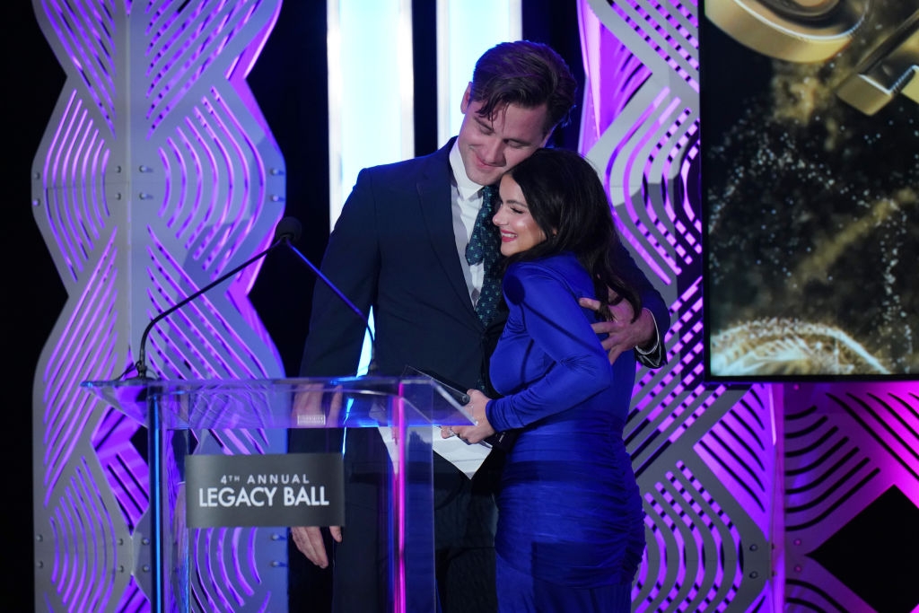 Two people embrace on stage at the Legacy Ball. The person on the left wears a suit, and the person on the right wears an elegant, fitted dress
