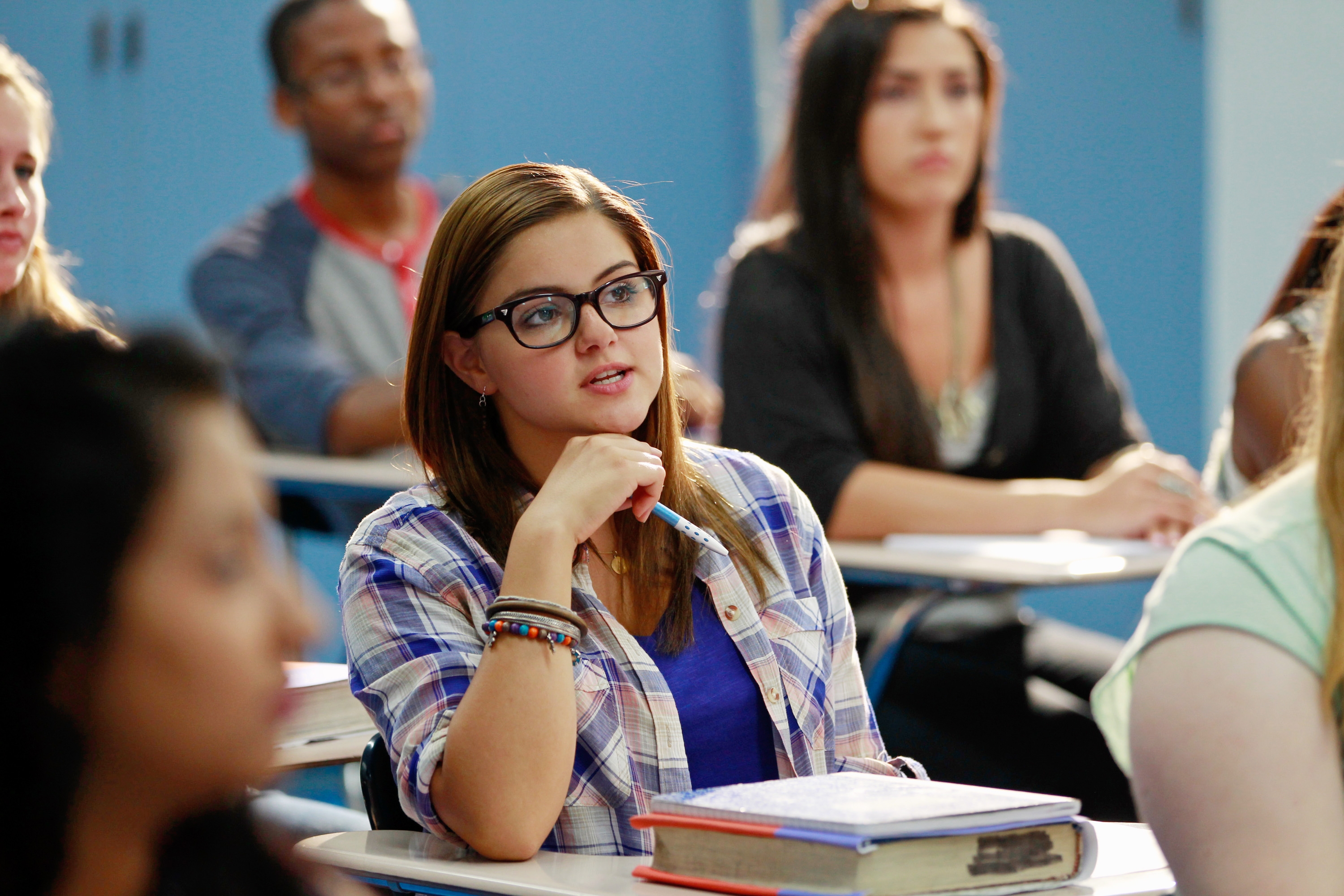 A seated student wearing glasses and a plaid shirt attentively listens in a classroom. Other students in blurred background
