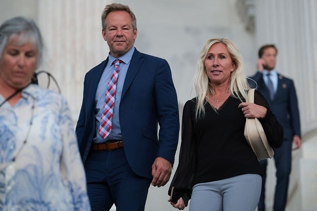 Two individuals in business attire walk together; the man wears a suit with a patterned tie, and the woman carries a bag. Others are in the background