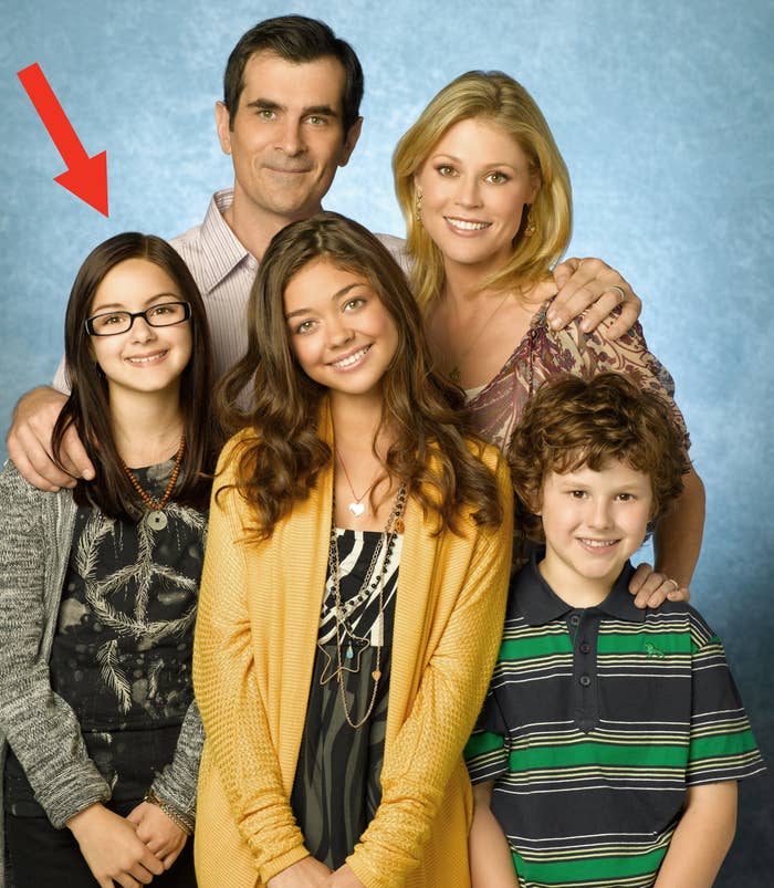 Five people smiling, with two adults standing behind three children. The group is posing formally against a plain background