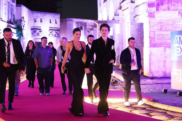 Two people in elegant black evening wear walking on a red carpet, flanked by others