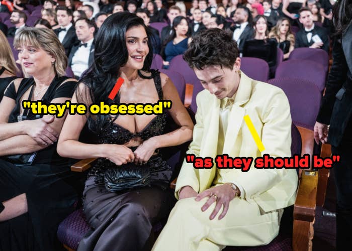 Two people seated at an event. One wears a sparkly dress, the other a light-colored suit. Audience visible in the background
