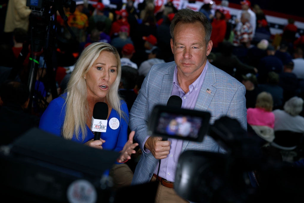 Two people speak into microphones at an event. The woman wears a buttoned outfit, and the man wears a blazer with a pin. Audience blurred in the background