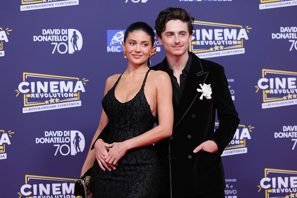 Two people pose on a red carpet; the woman wears a sleeveless lace dress, and the man is in a velvet suit with floral detail