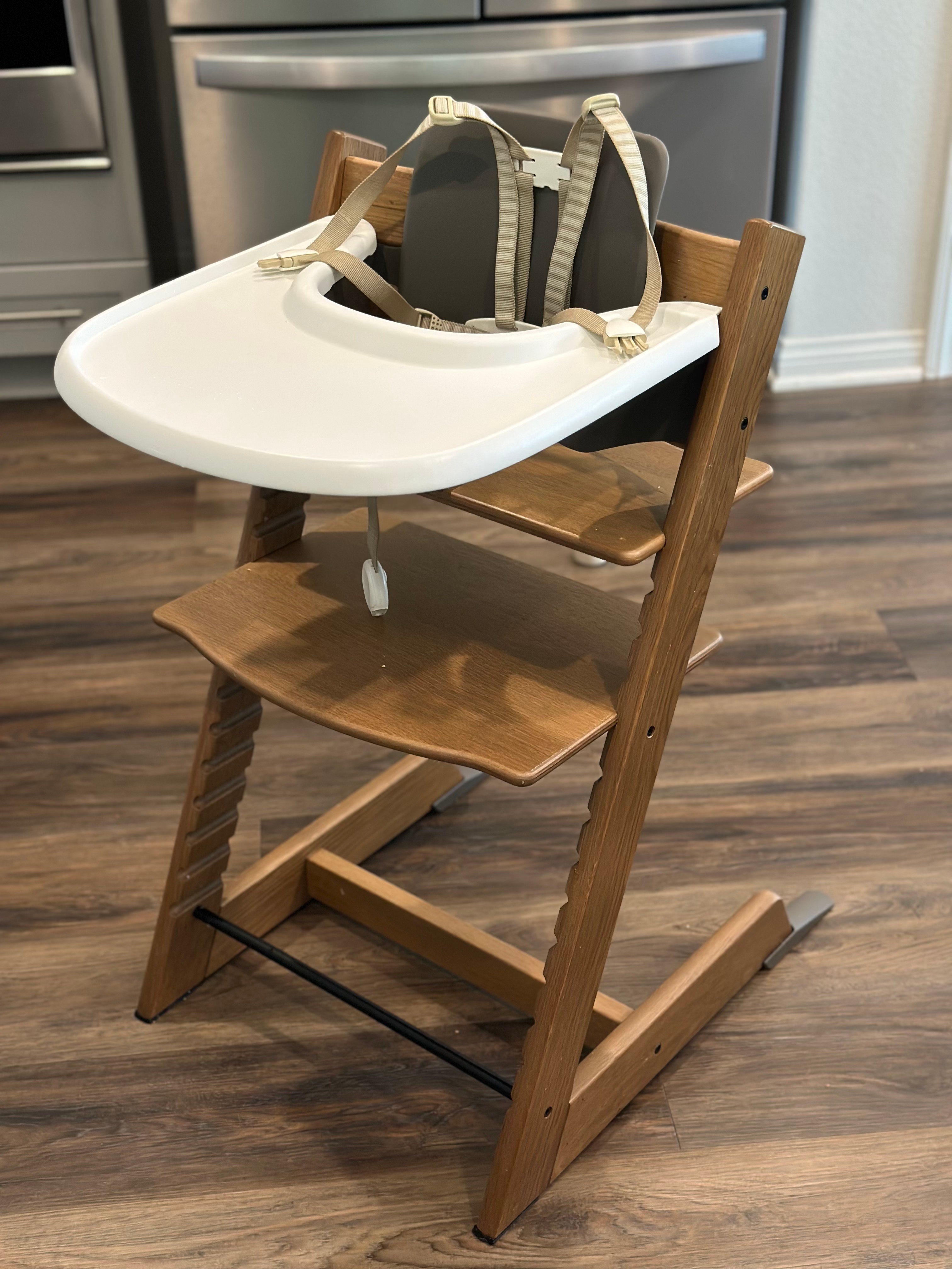 Wooden high chair with a white tray and adjustable strap, set on a wooden floor in a kitchen setting