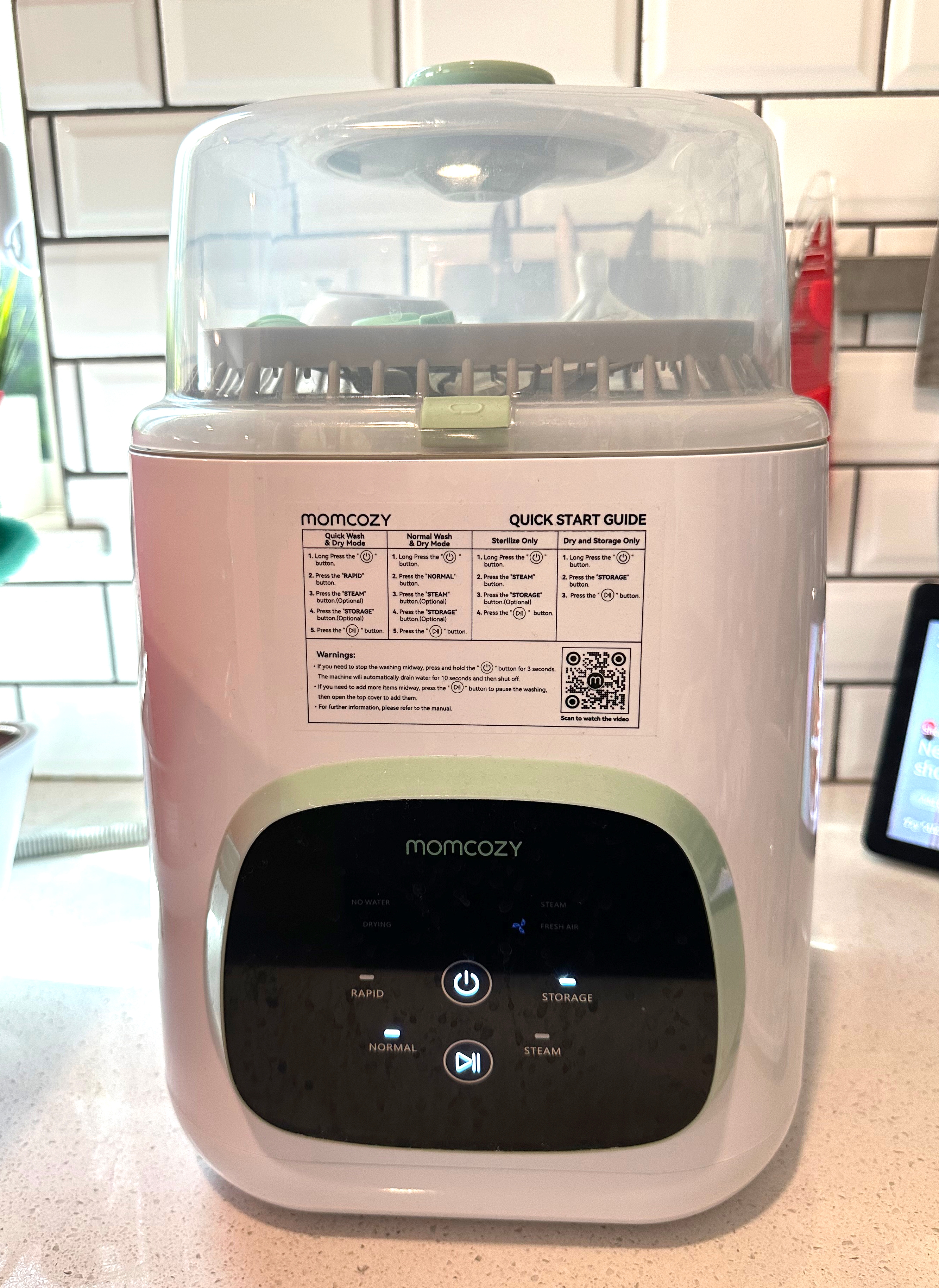 Baby bottle sterilizer on a kitchen counter with label showing instructions and a QR code