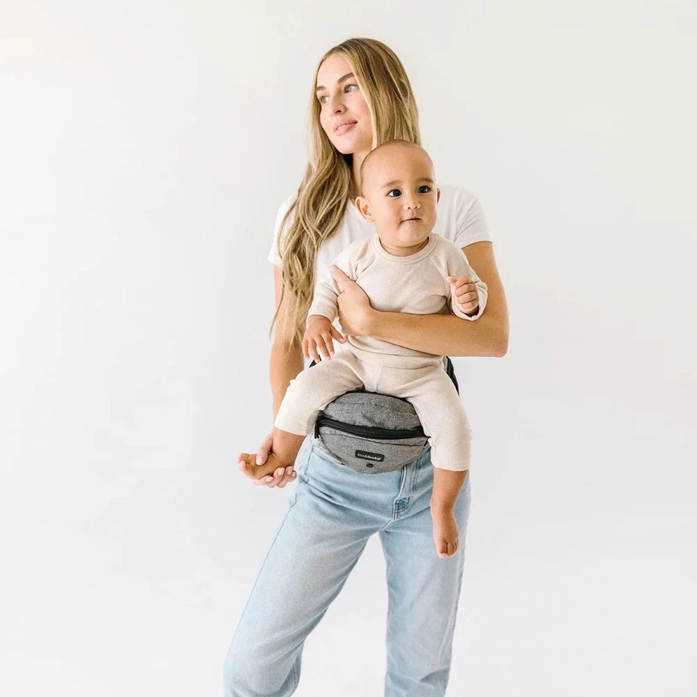 Woman with long hair holding a baby on a hip seat. Both are in casual attire, standing against a plain background