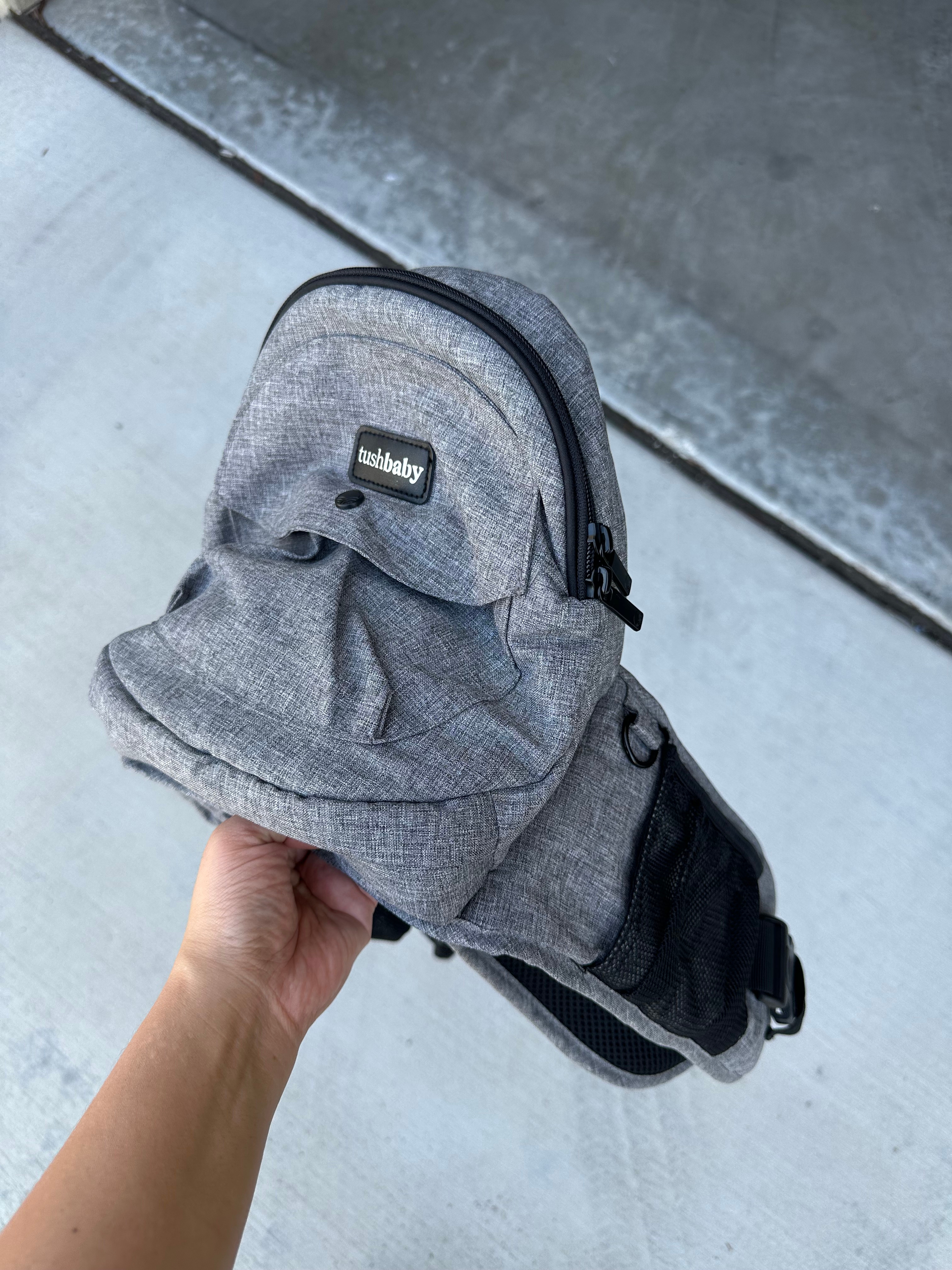 Hand holding a gray baby carrier with a label reading "tushbaby," featuring pockets and a strap