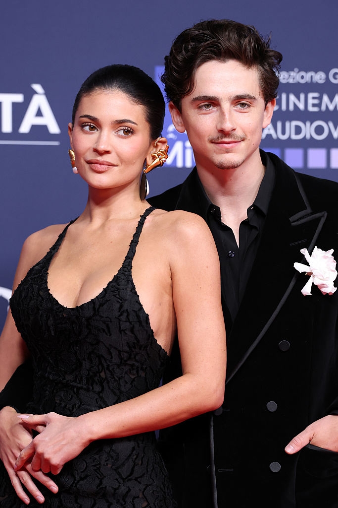 Two individuals on a red carpet; one wearing a sleeveless textured gown, the other in a velvet suit with a flower accessory