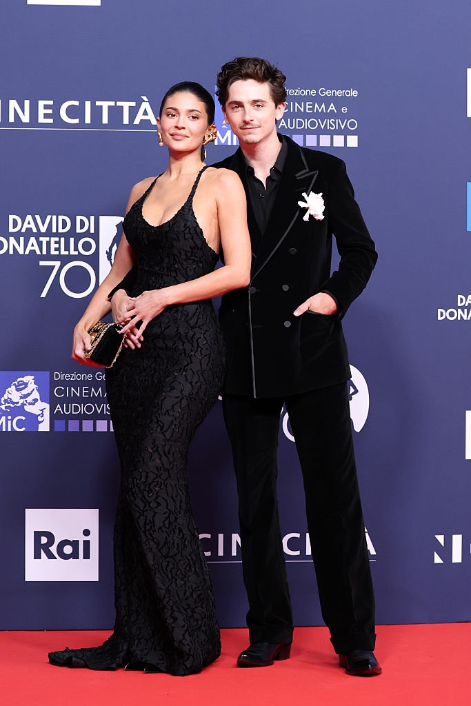 Two people pose on a red carpet. One wears a black halter gown; the other wears a black suit with a white boutonniere