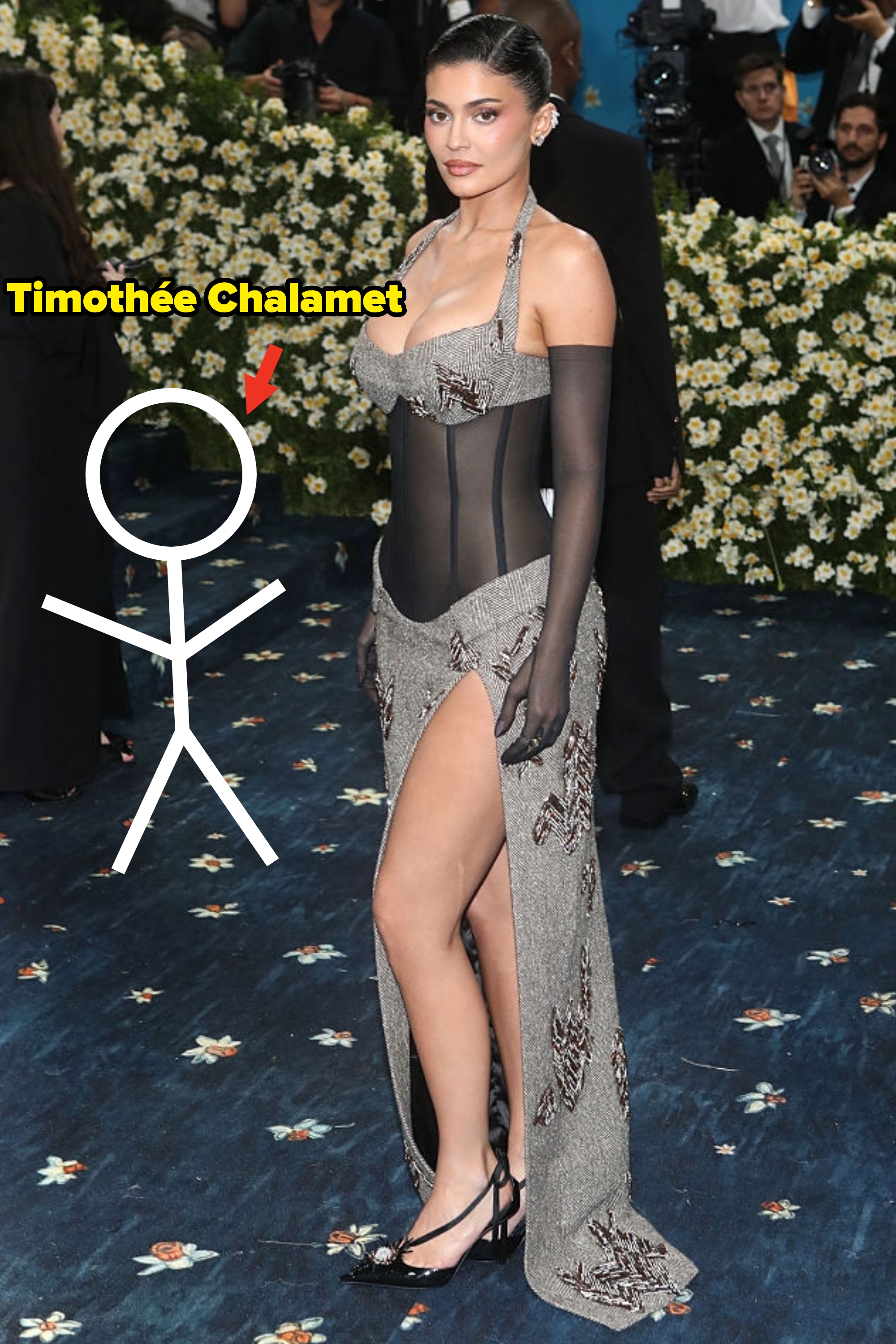 Person on a red carpet wearing a form-fitting, sheer gown with ornate detailing and high slit, surrounded by photographers and floral decorations