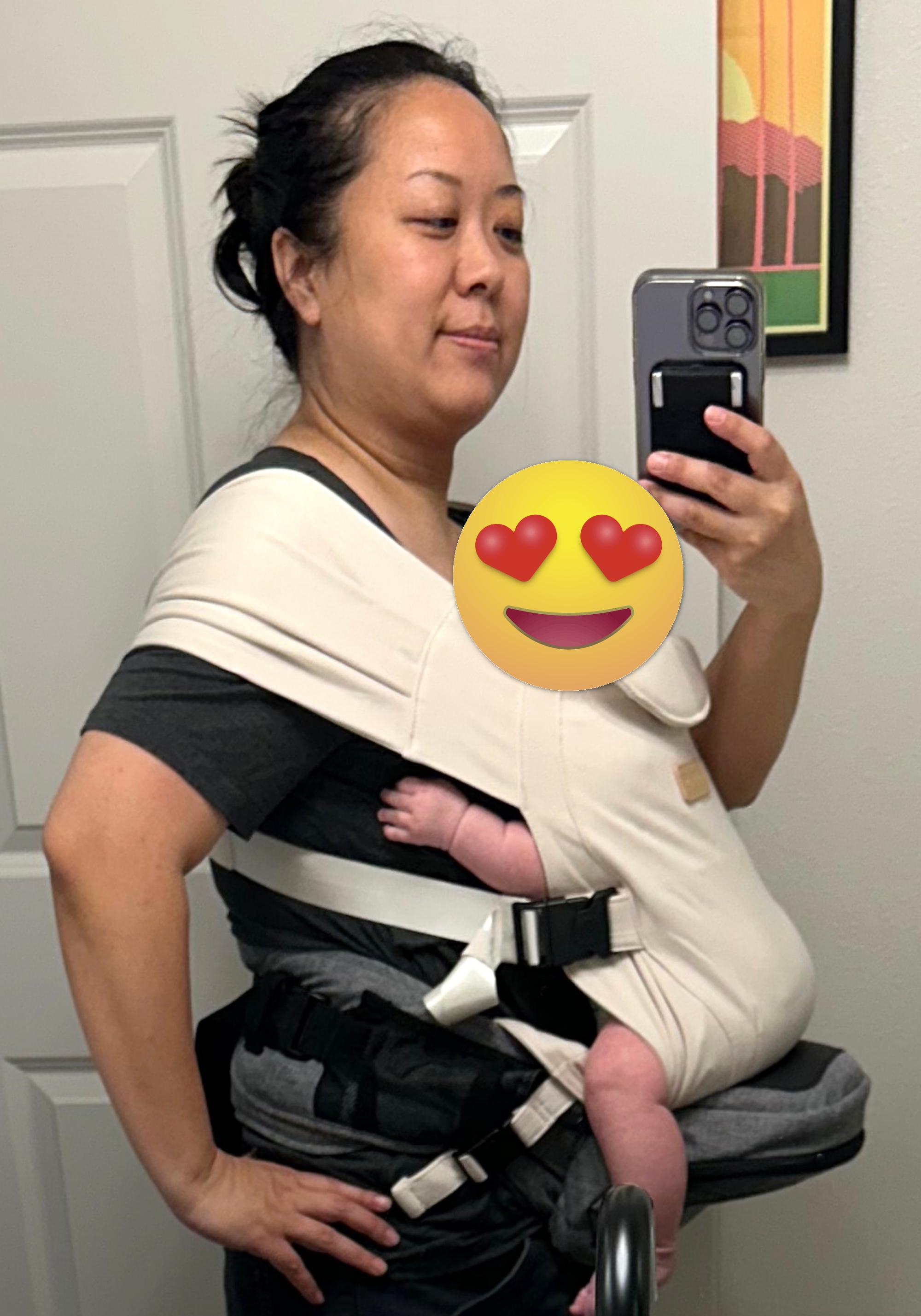 A person takes a mirror selfie with a baby secured in a soft baby carrier