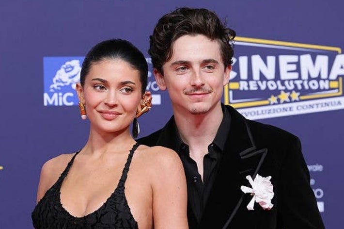 A man and woman pose on the red carpet, both in stylish evening wear. The woman wears a dress with a textured bodice, and the man an embroidered suit