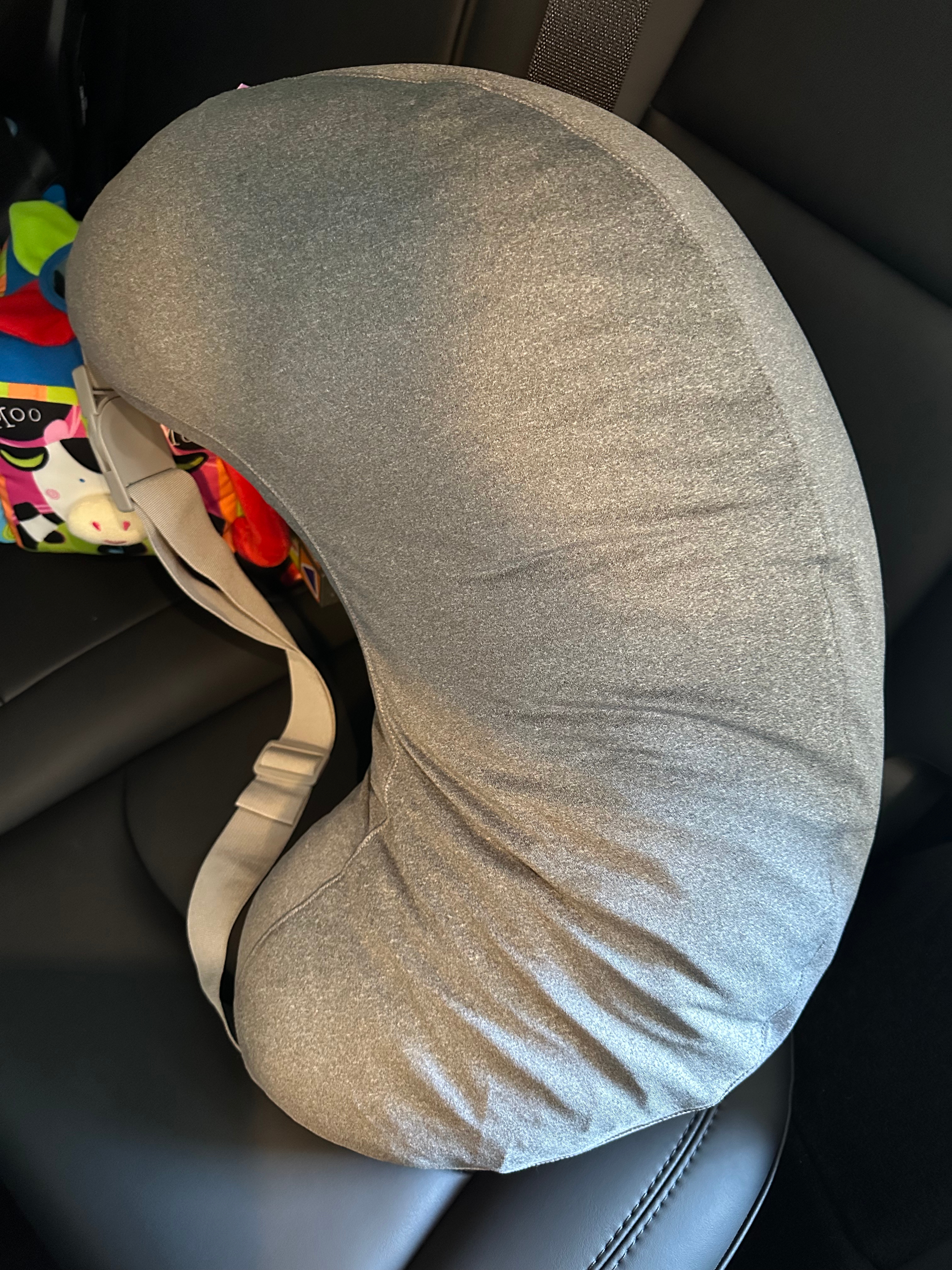 Half-moon shaped nursing pillow with a gray fabric cover, resting on a black car seat, with a colorful bag in the background
