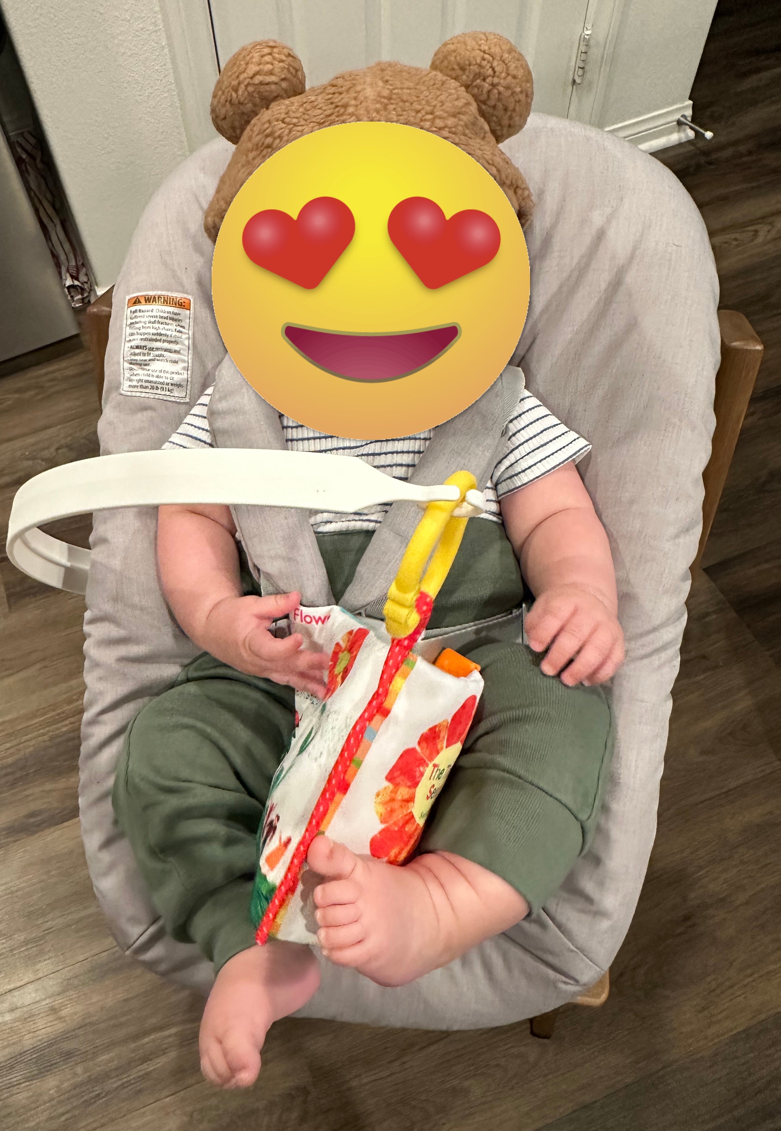 Baby in bear-eared hat sits in a chair, wearing a striped shirt and green pants, holding a colorful packaged snack
