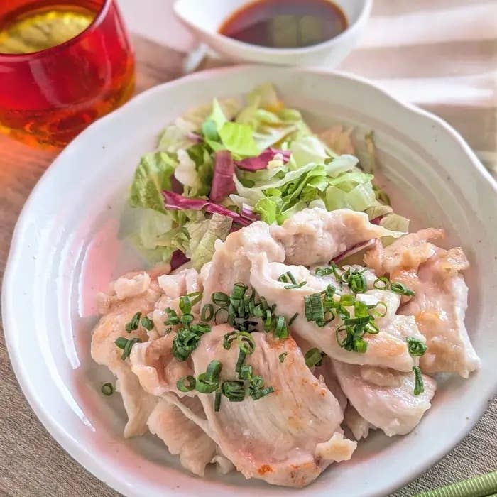 お皿に盛り付けられたゆでた鶏肉と刻みネギ、生野菜のサラダが添えられています。タレがそばにあります。