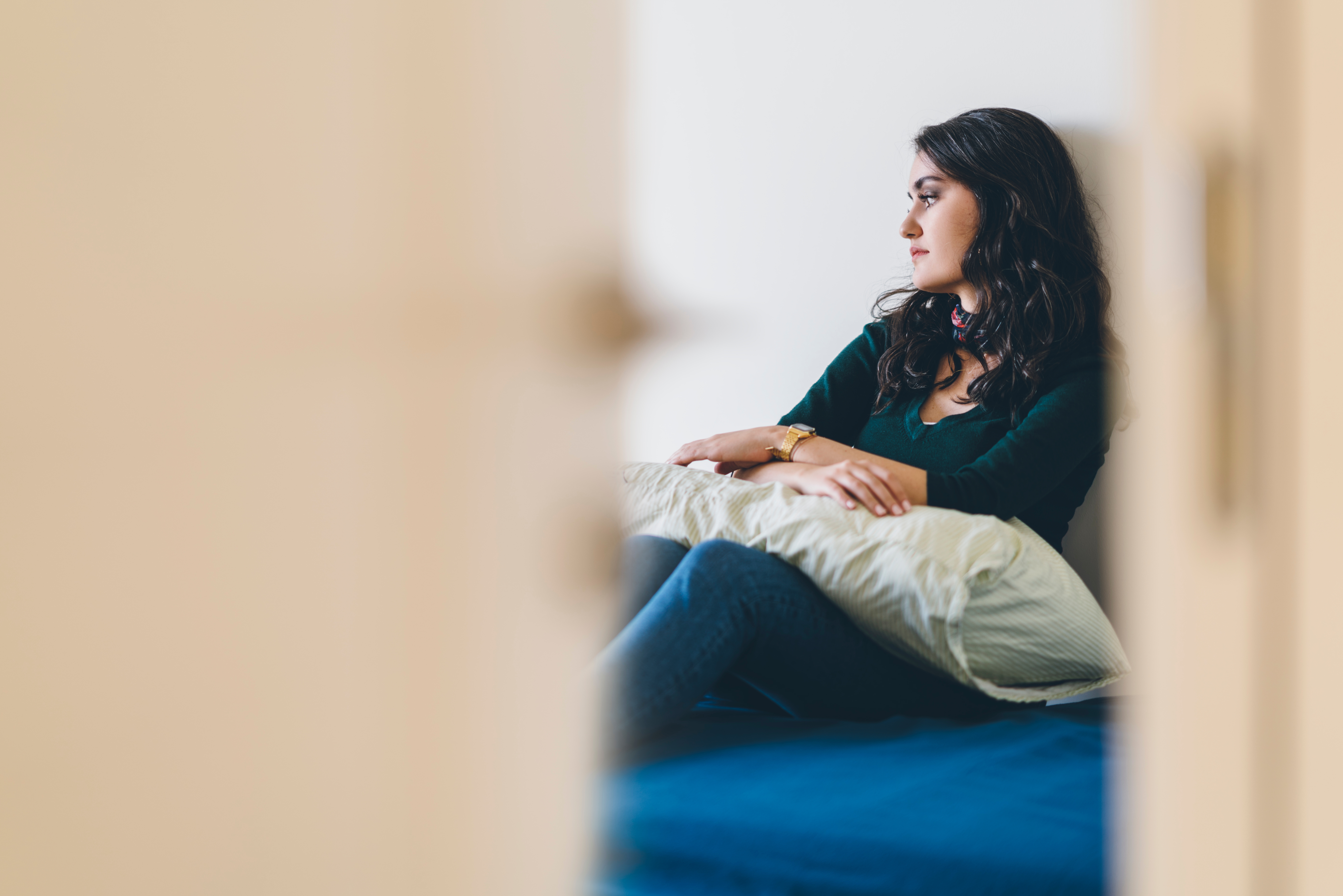 A woman sits pensively on a bed, holding a pillow and looking out of view. A door frames the scene, adding a sense of privacy