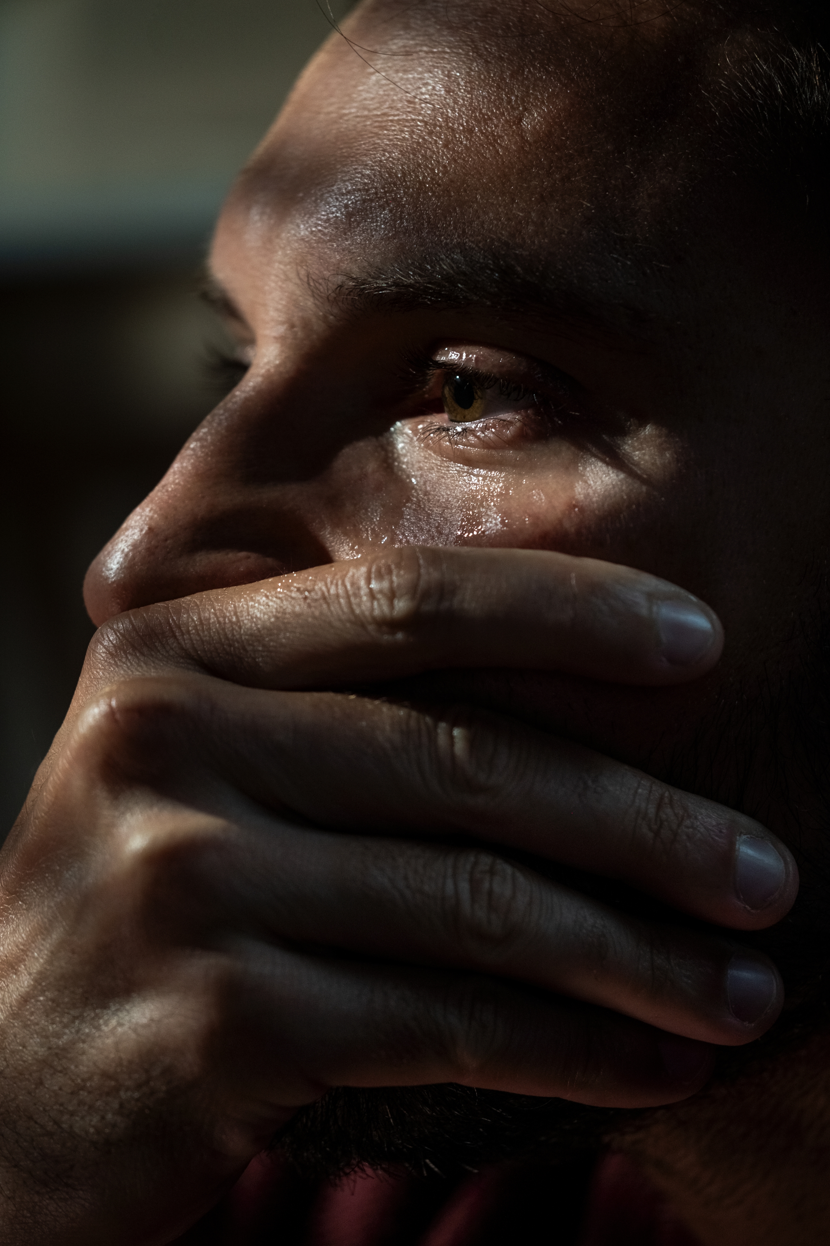 Close-up of a person with a contemplative expression, hand covering their mouth, light casting shadows on their face