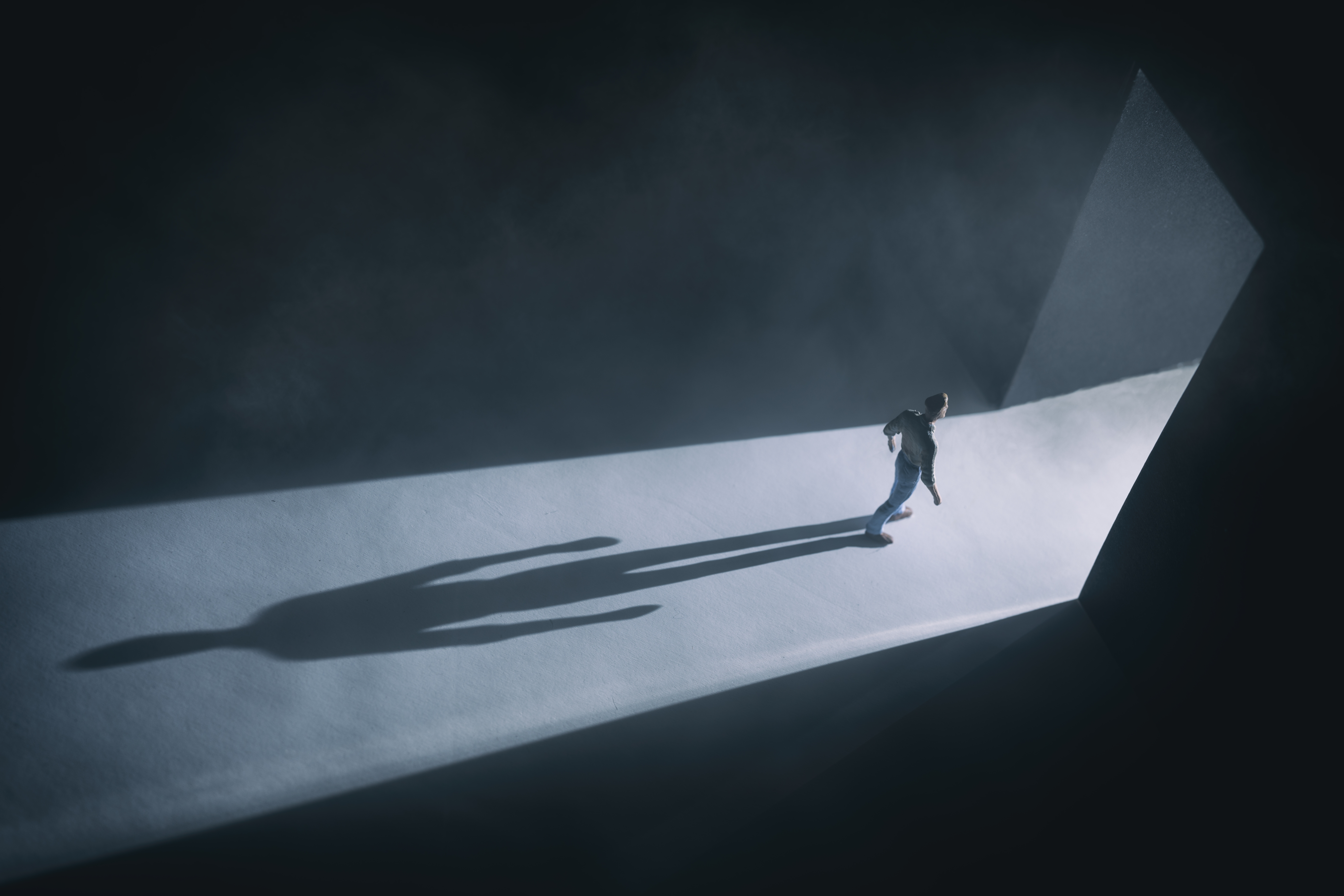 A person stands dramatically under a spotlight in a dark, misty room, casting a long shadow toward an open doorway
