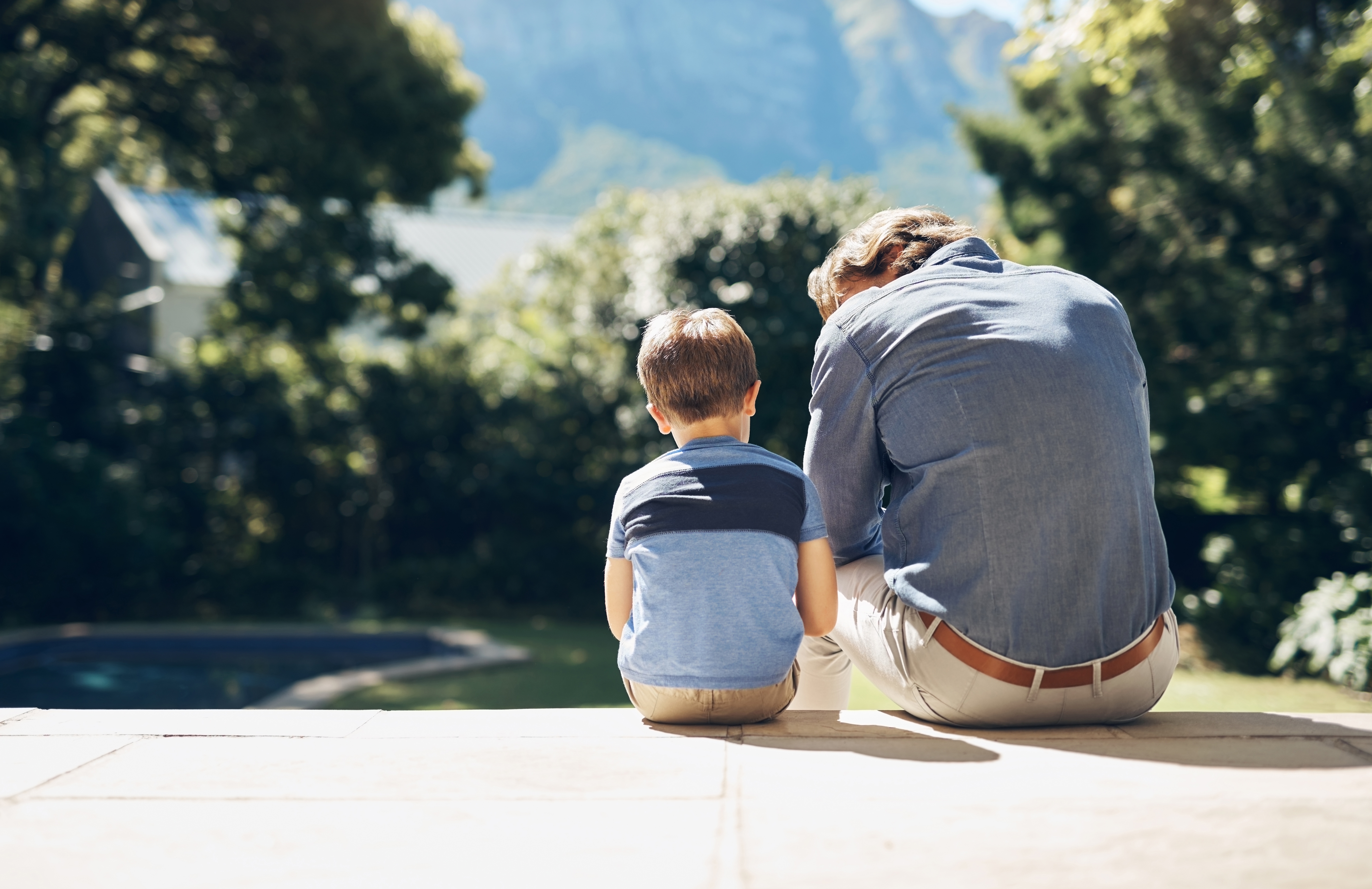 An adult and child sit together on a step, facing a scenic view with trees and mountains, sharing a quiet moment