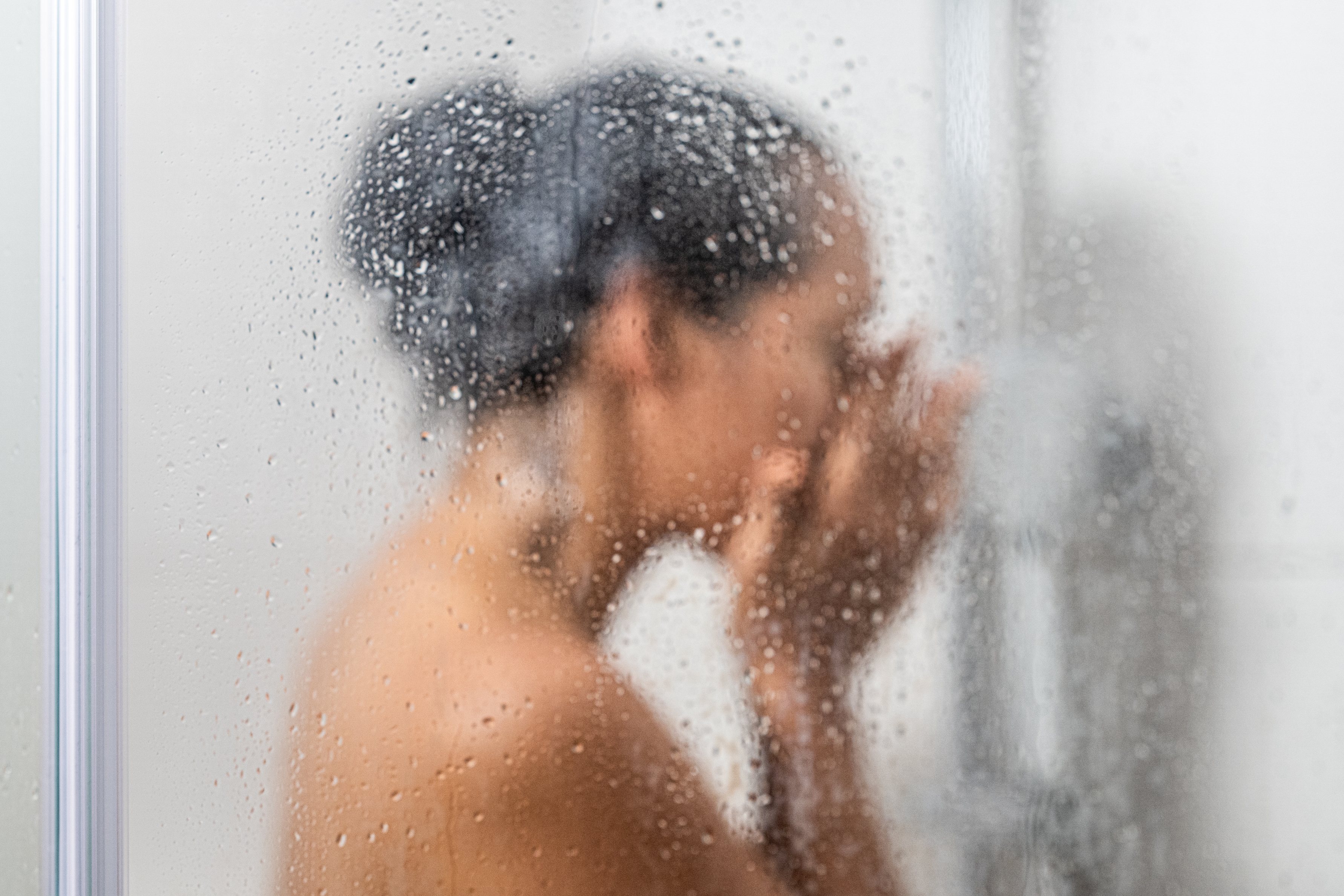Person in a shower behind a steamed glass door, gently touching their face, creating a calm and intimate atmosphere