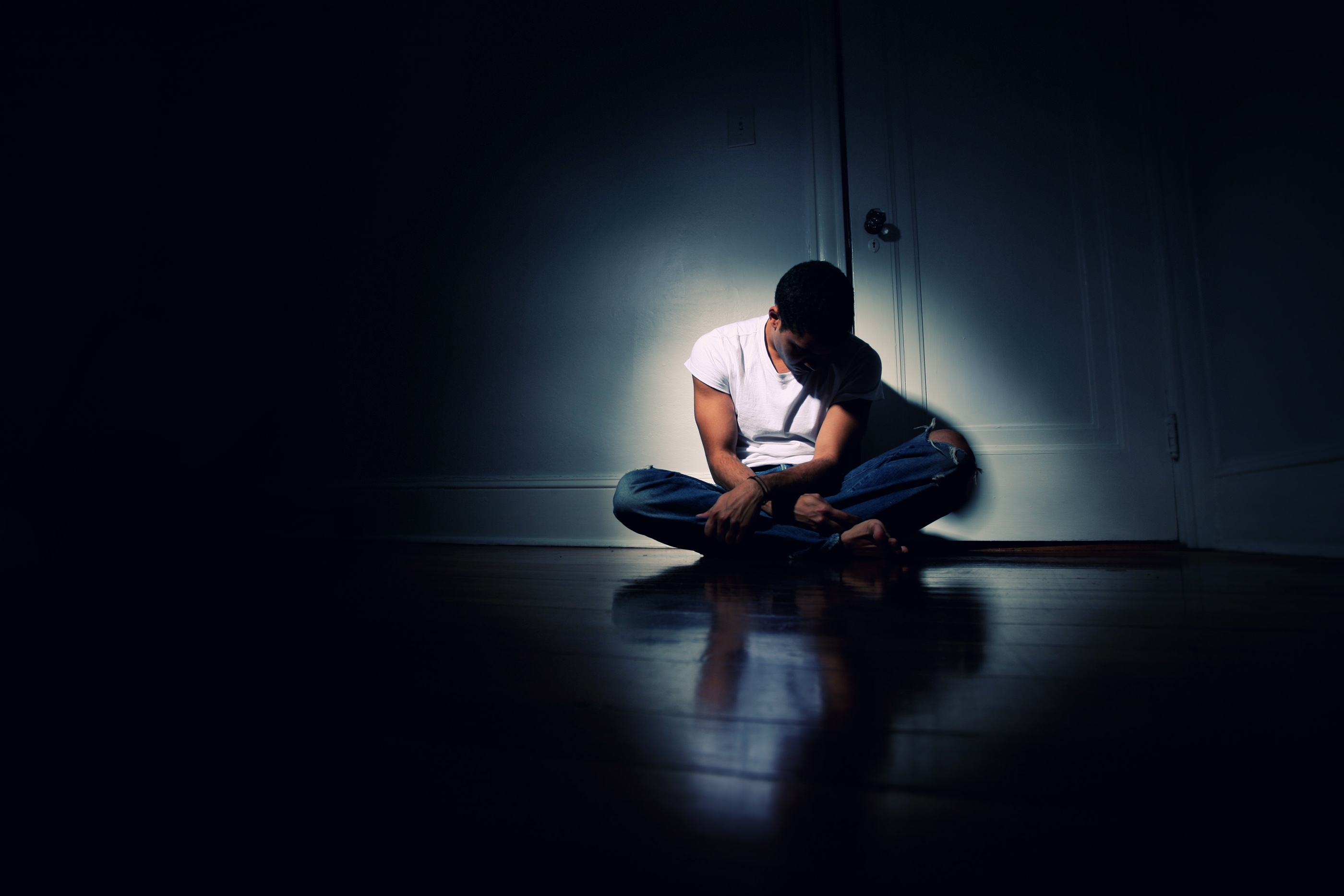Person sitting on the floor in a dimly lit room, head down, wearing a T-shirt and jeans. Light focuses on them, accentuating the solitary mood