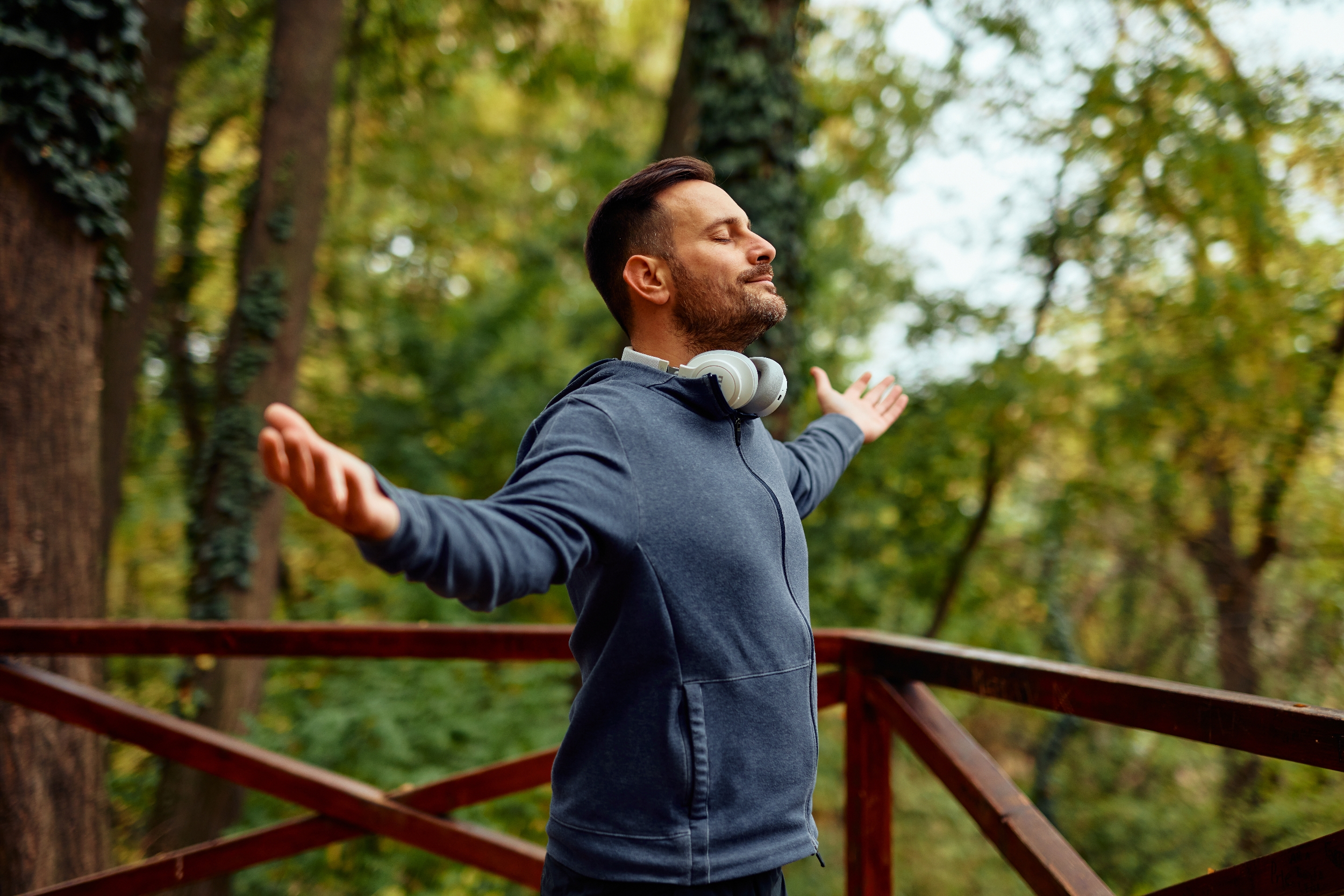 Man in casual hoodie stands on a wooden deck with arms outstretched, enjoying the serene forest surroundings