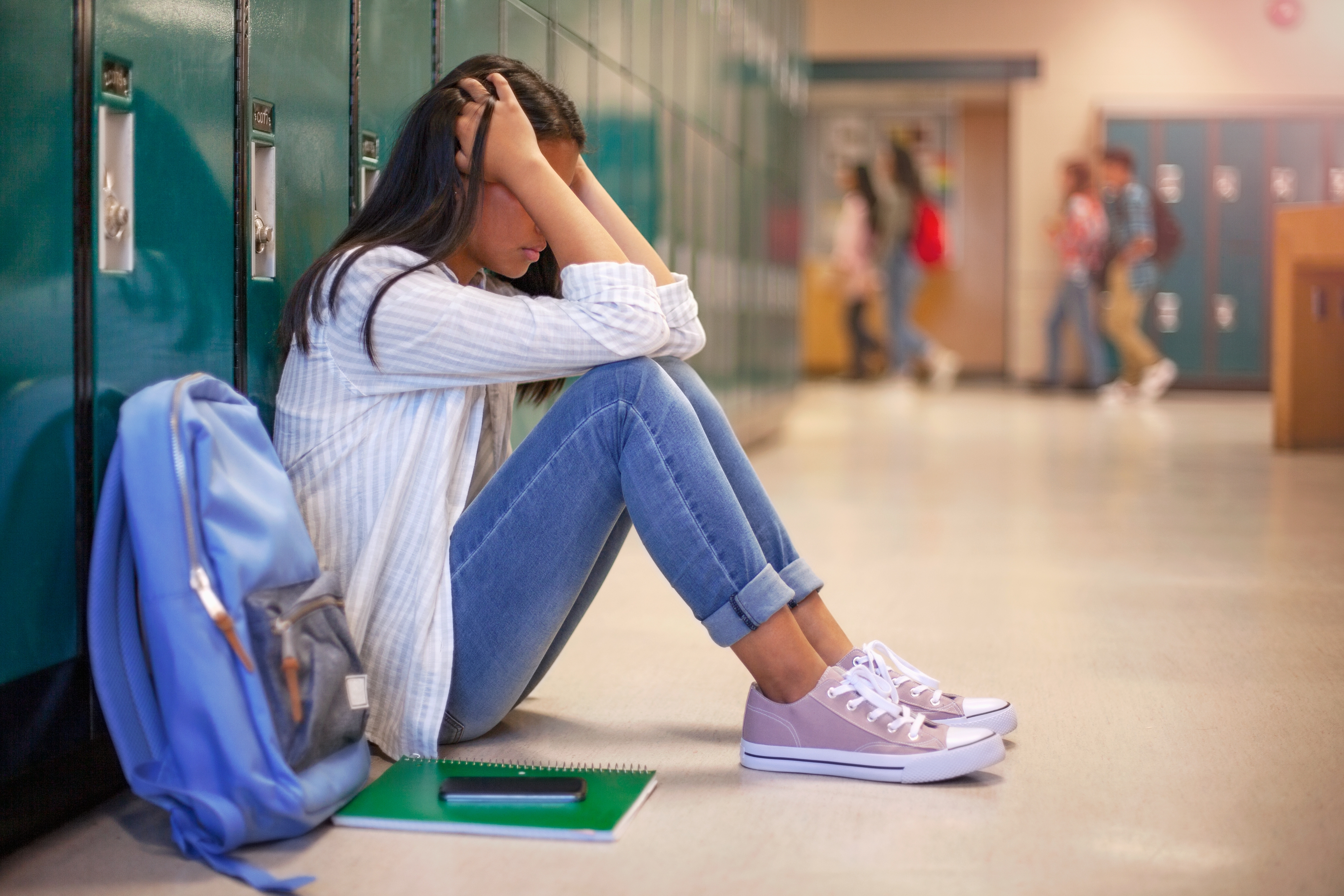 Student sitting against lockers, head in hands, looking distressed. Notebook and backpack on the floor beside them. Other students are in the background
