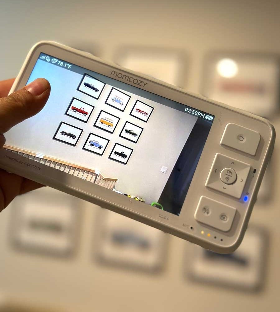 A hand holds a baby monitor showing a crib and framed car pictures on the wall