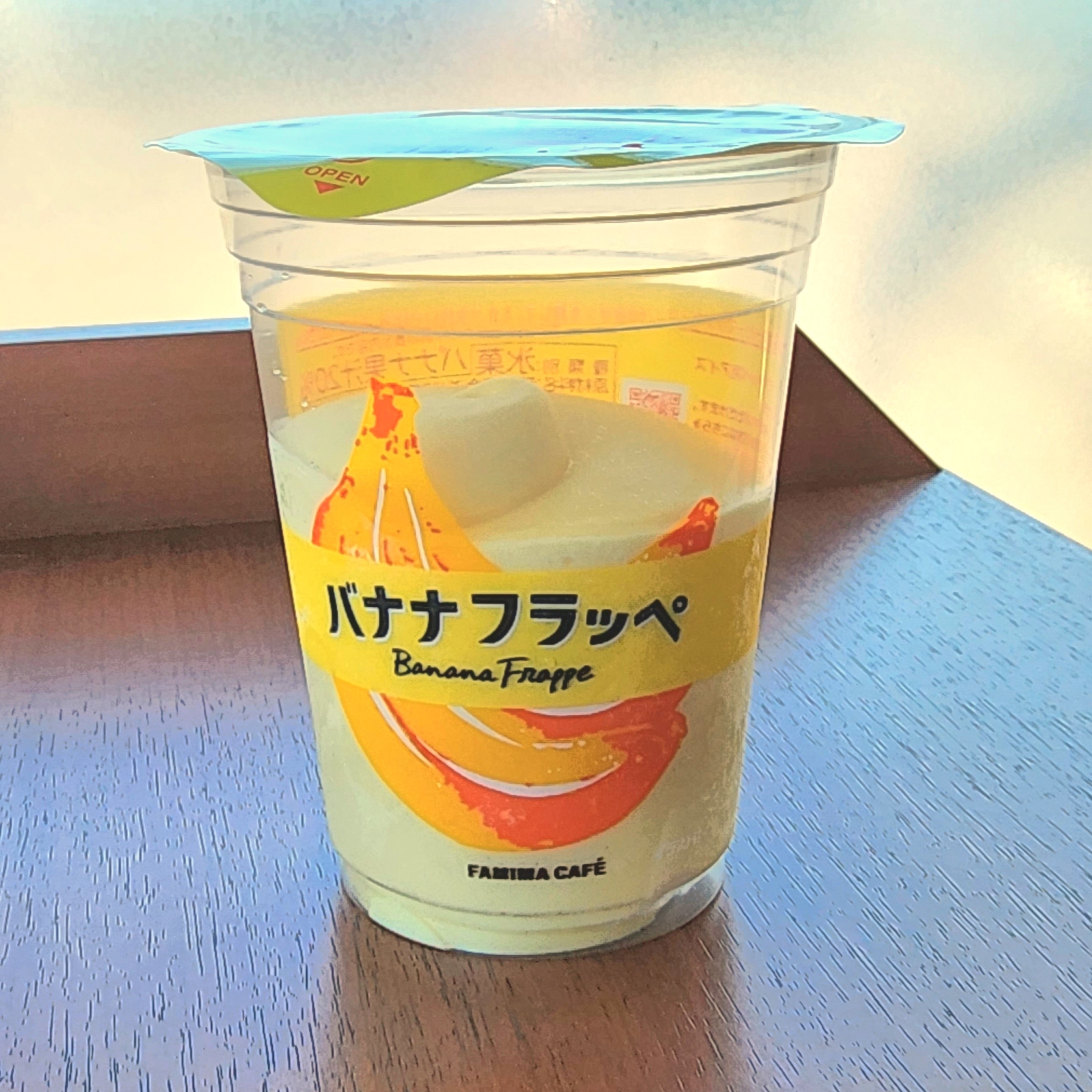 ファミマカフェのバナナフラッペのカップが木製テーブルに置かれている。