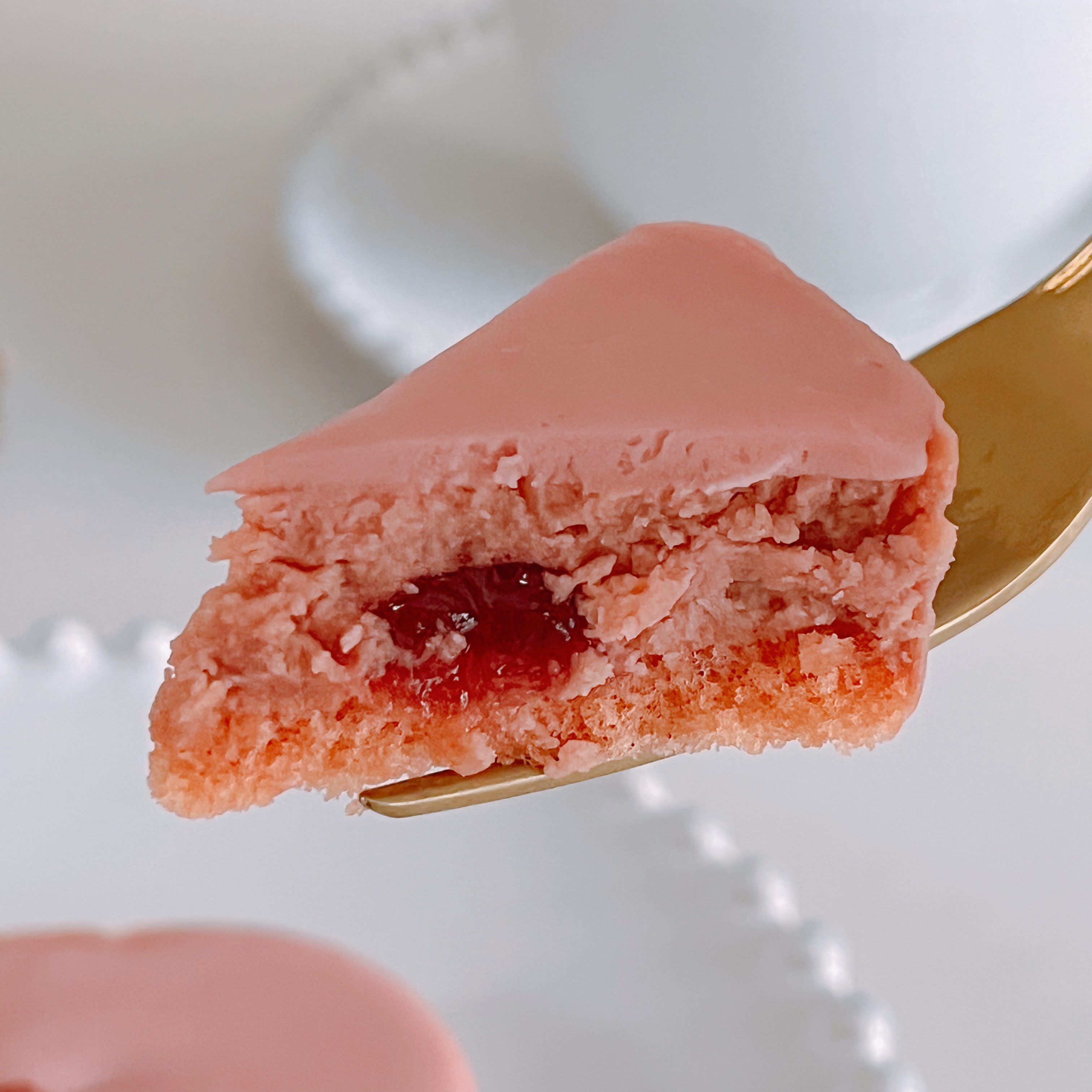 フォークにのったピンクのマカロン、中心に赤いジャムが見える。背景にぼんやりとした食器がある。