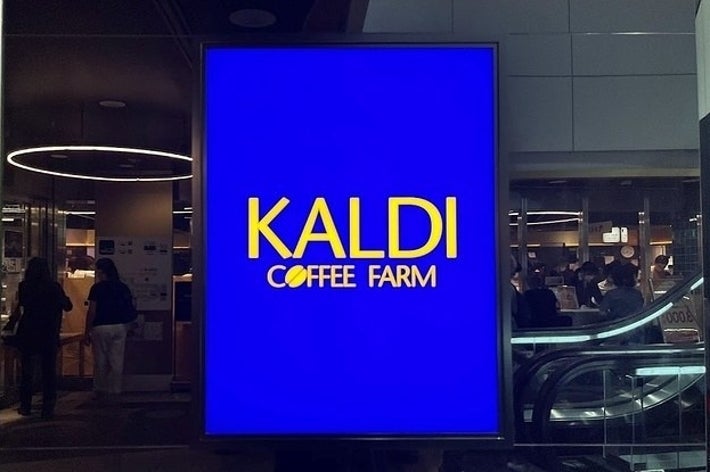 カフェ「カルディコーヒーファーム」の大きな店頭看板。背景の店舗には買い物客が見える。