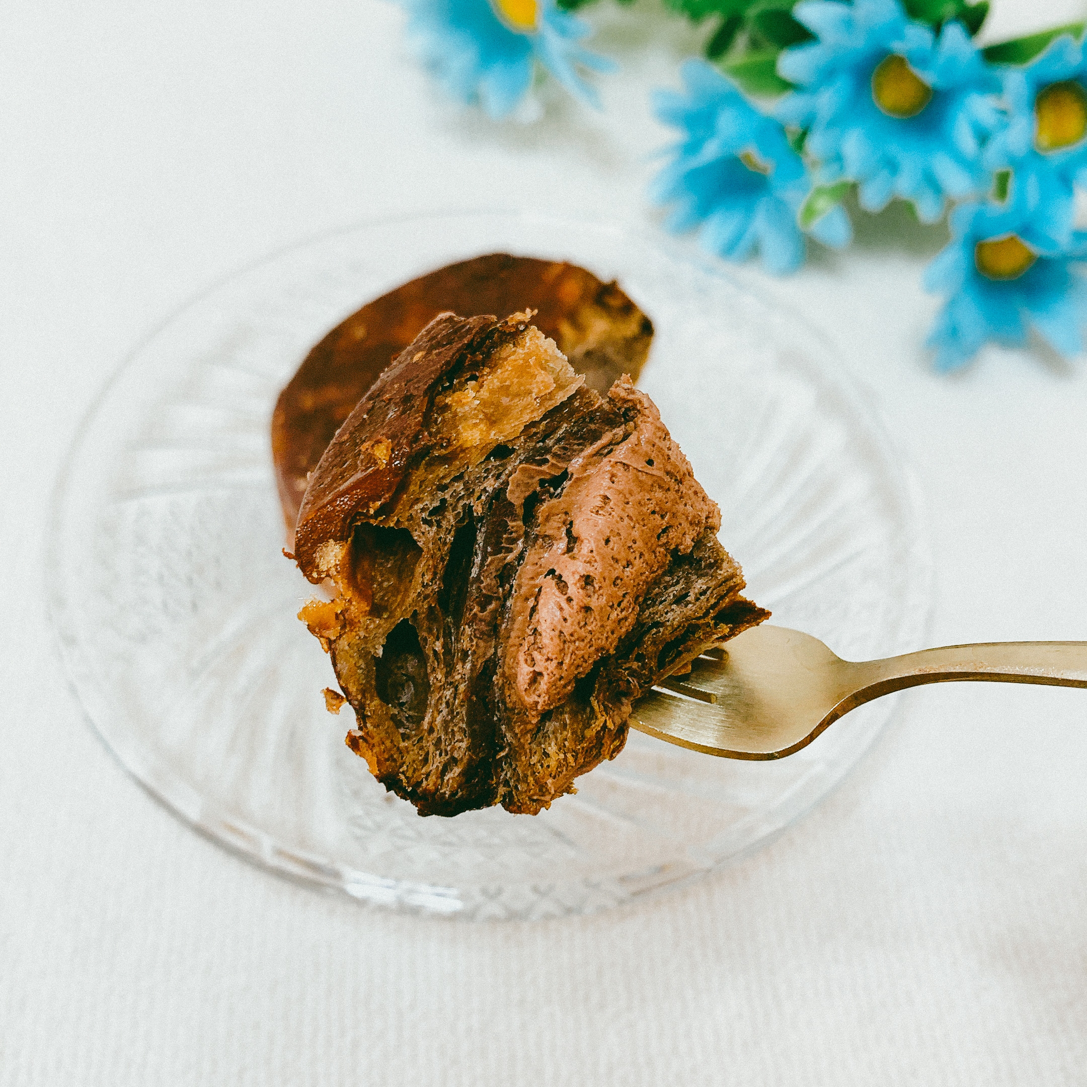 フォークに刺さったチョコレートケーキの一部。背景に青い花が添えられたガラスの皿が見える。