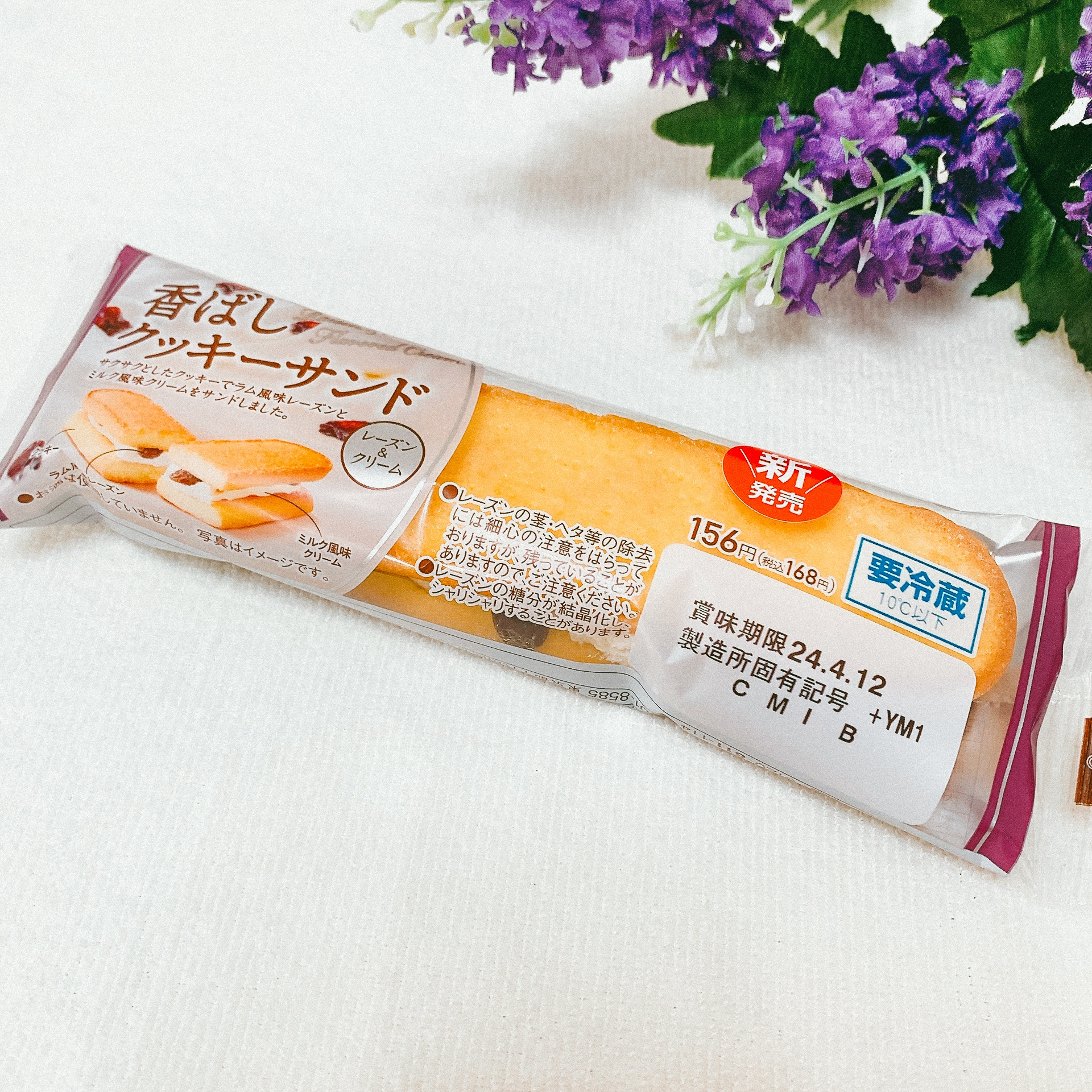 「香ばしクッキーサンド」と書かれたパッケージに包まれたお菓子がテーブルに置かれている。横に紫の花がある。