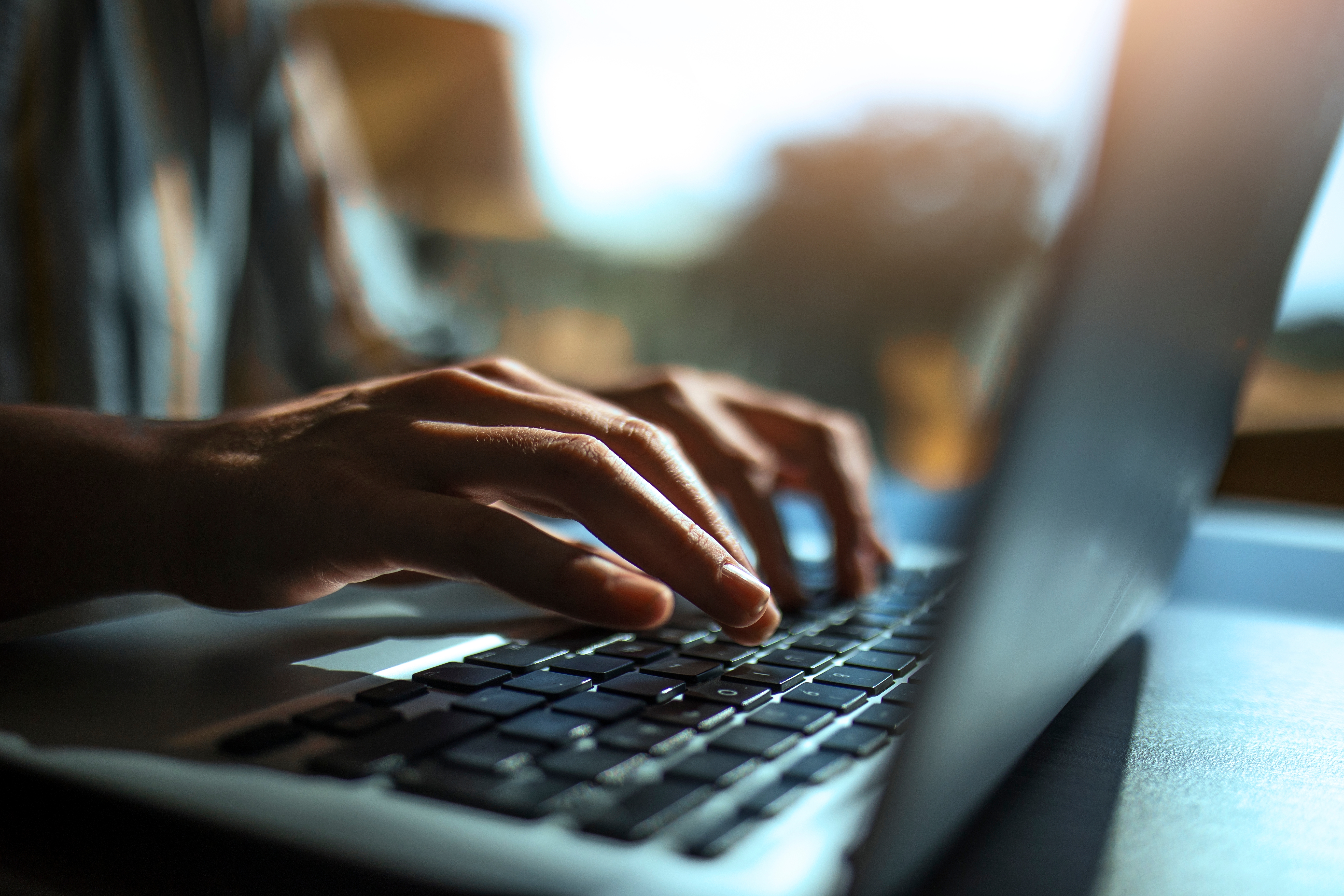 A person's hands typing on a laptop keyboard, with natural lighting in the background.