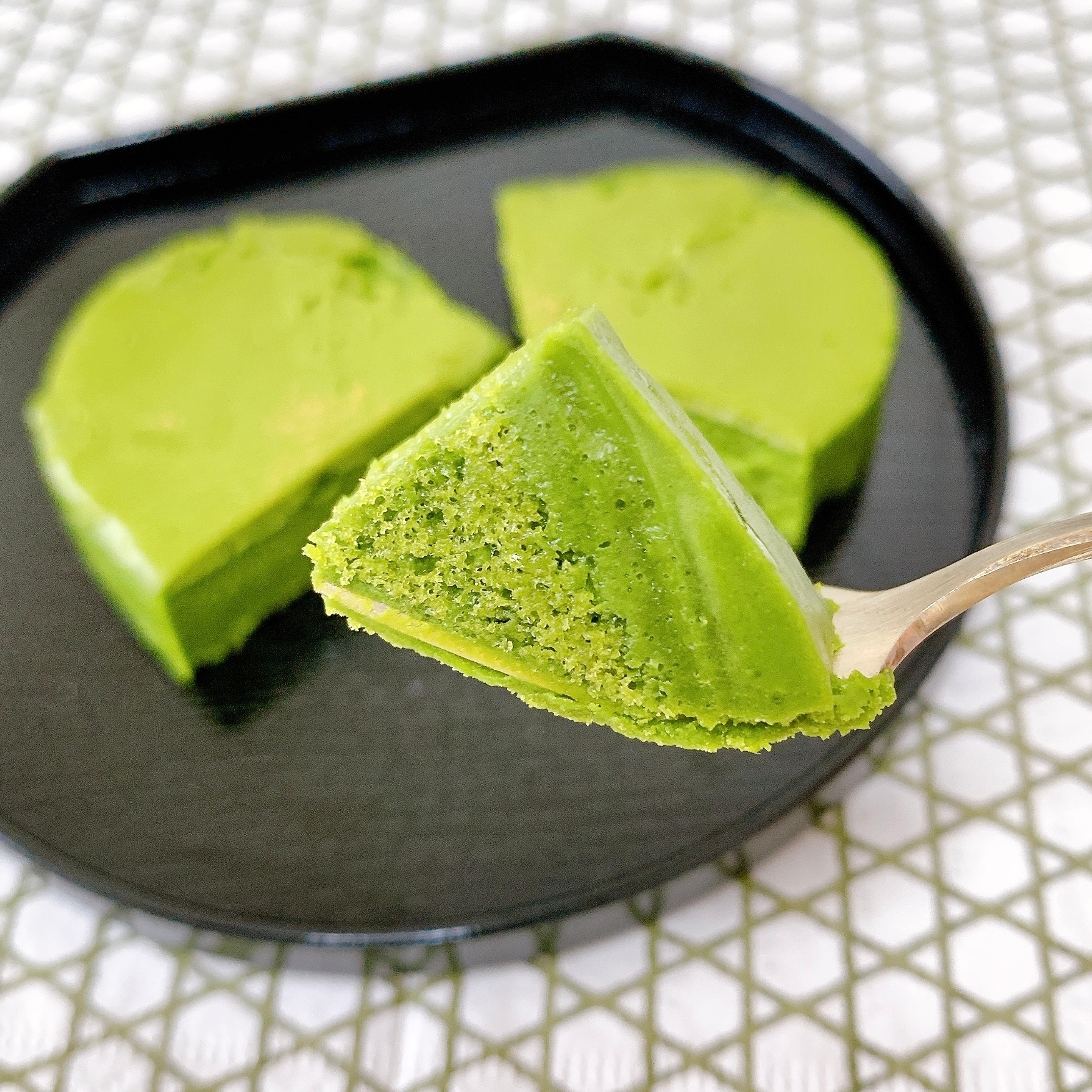 フォークにのった抹茶ケーキが黒い皿の上に盛られ、背景はチェック柄のテーブルクロス。