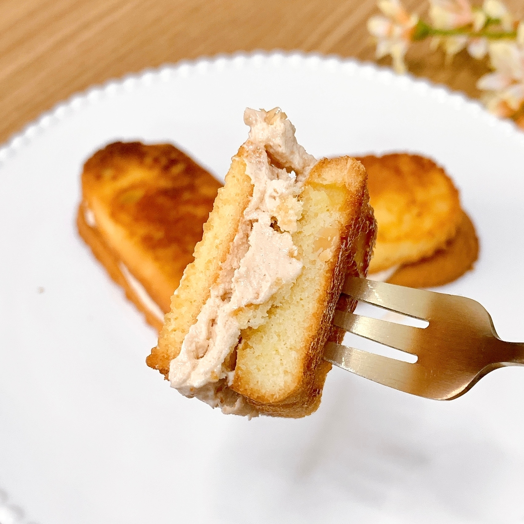 フォークに刺さったクリームの入ったケーキが白い皿に乗っている様子。背景に花が見える。