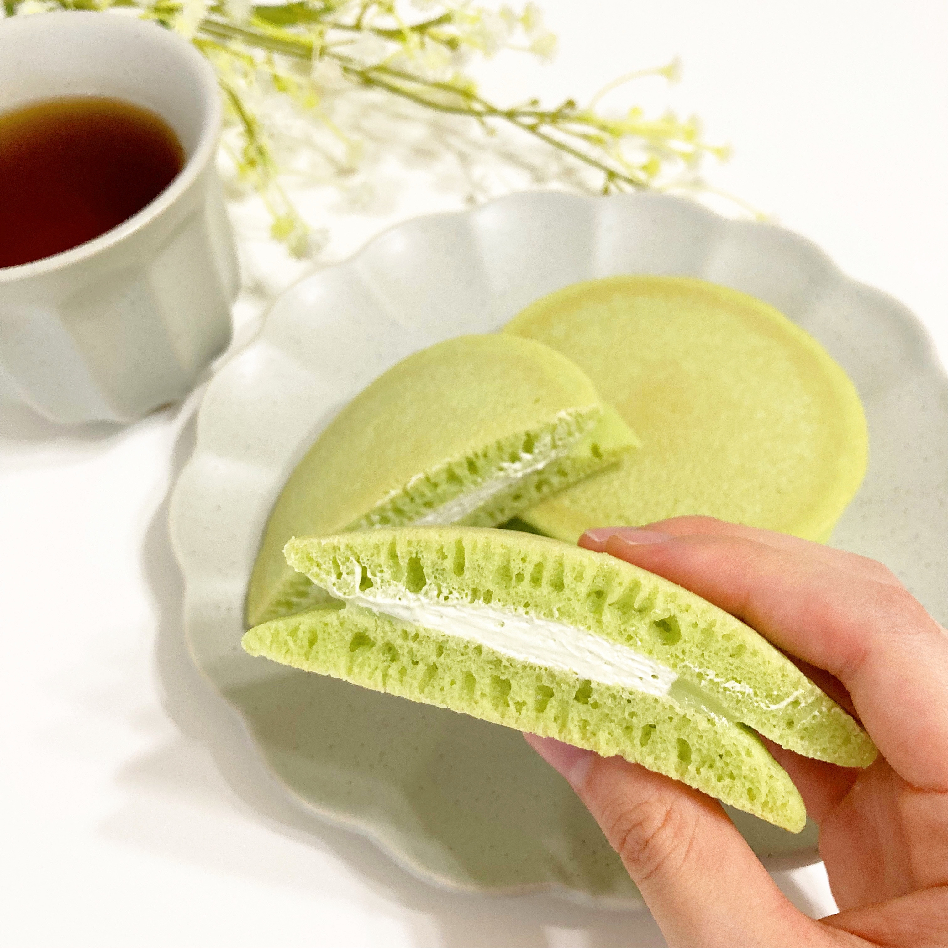 抹茶クリームサンドのパンケーキを手に持つ。後ろにコーヒーカップと白い花が飾られている。