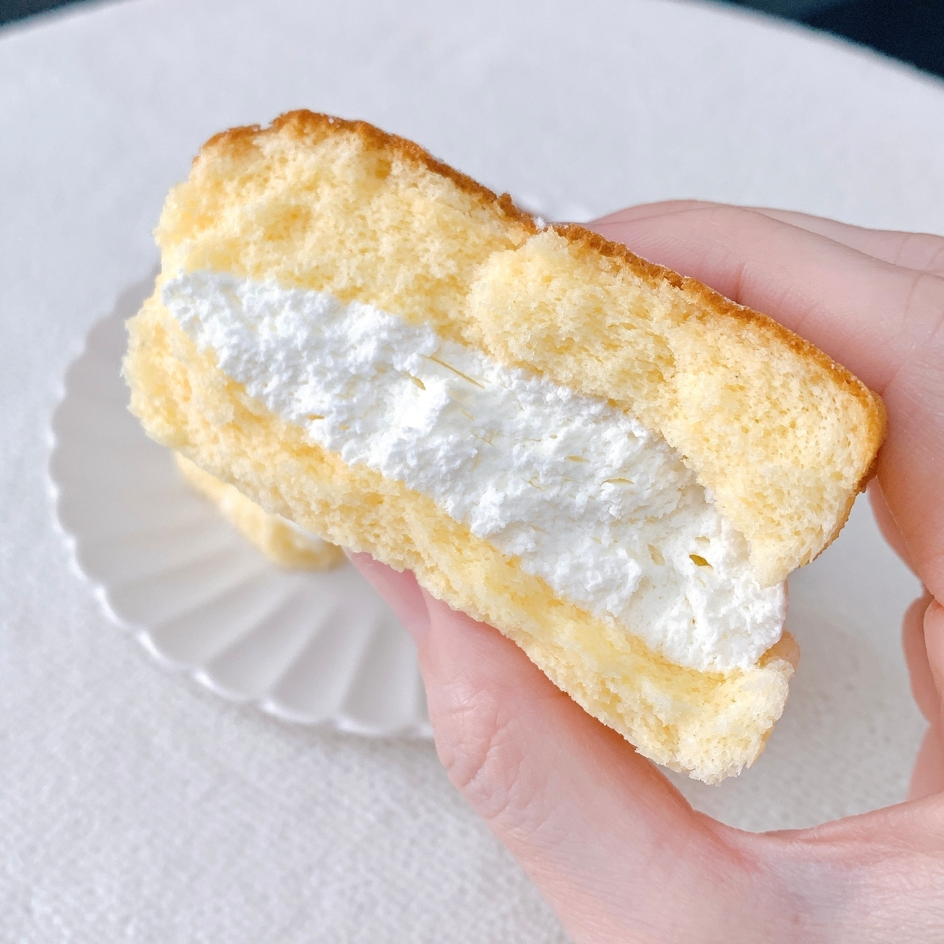 手に持たれたクリームたっぷりのふわふわサンドイッチケーキ。白い皿の上に載せられている。