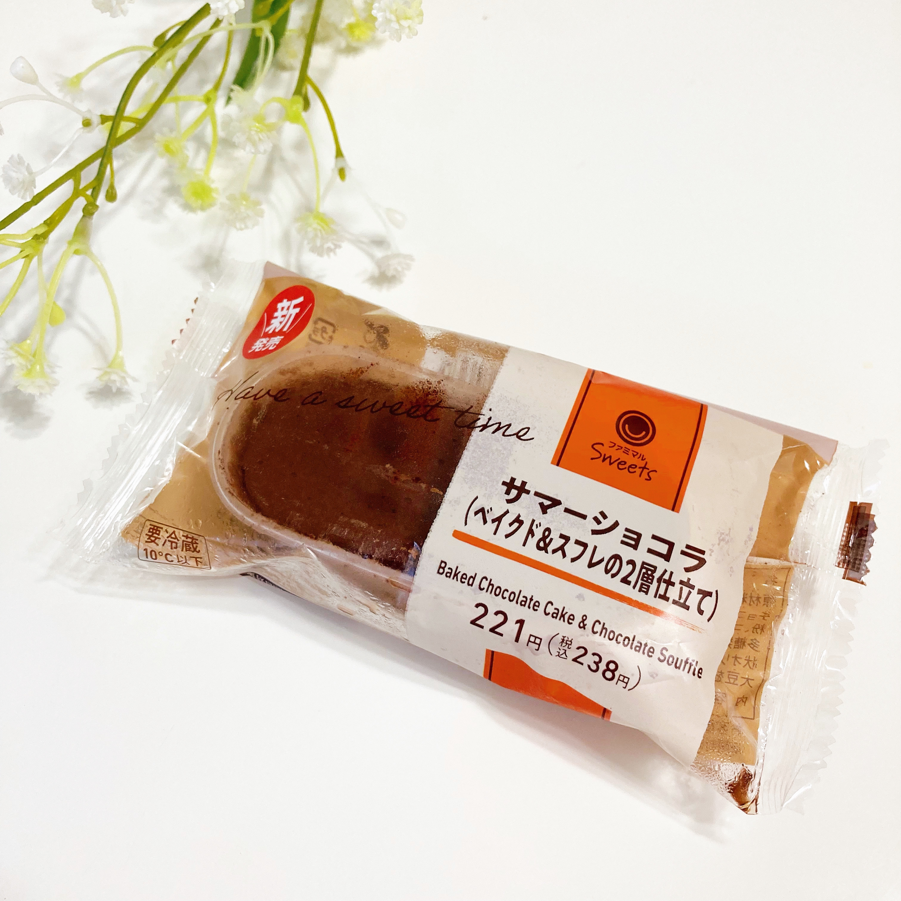 サマーショコラの包装されたチョコレートケーキが白い背景に置かれている。背景に小さな白い花も見える。