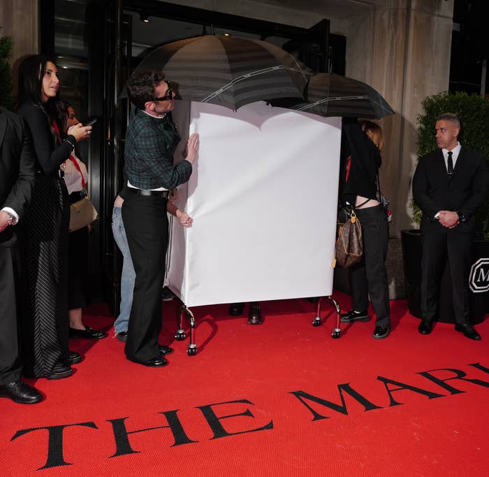 Doechii shielded by umbrellas and a white sheet on a red carpet at The Mark entrance, surrounded by security and photographers