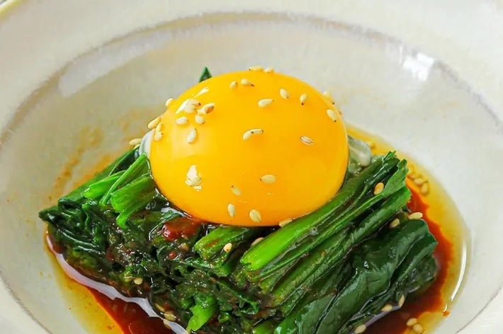 皿の上に盛り付けられた青菜と卵黄、ゴマとタレがかかっている料理。