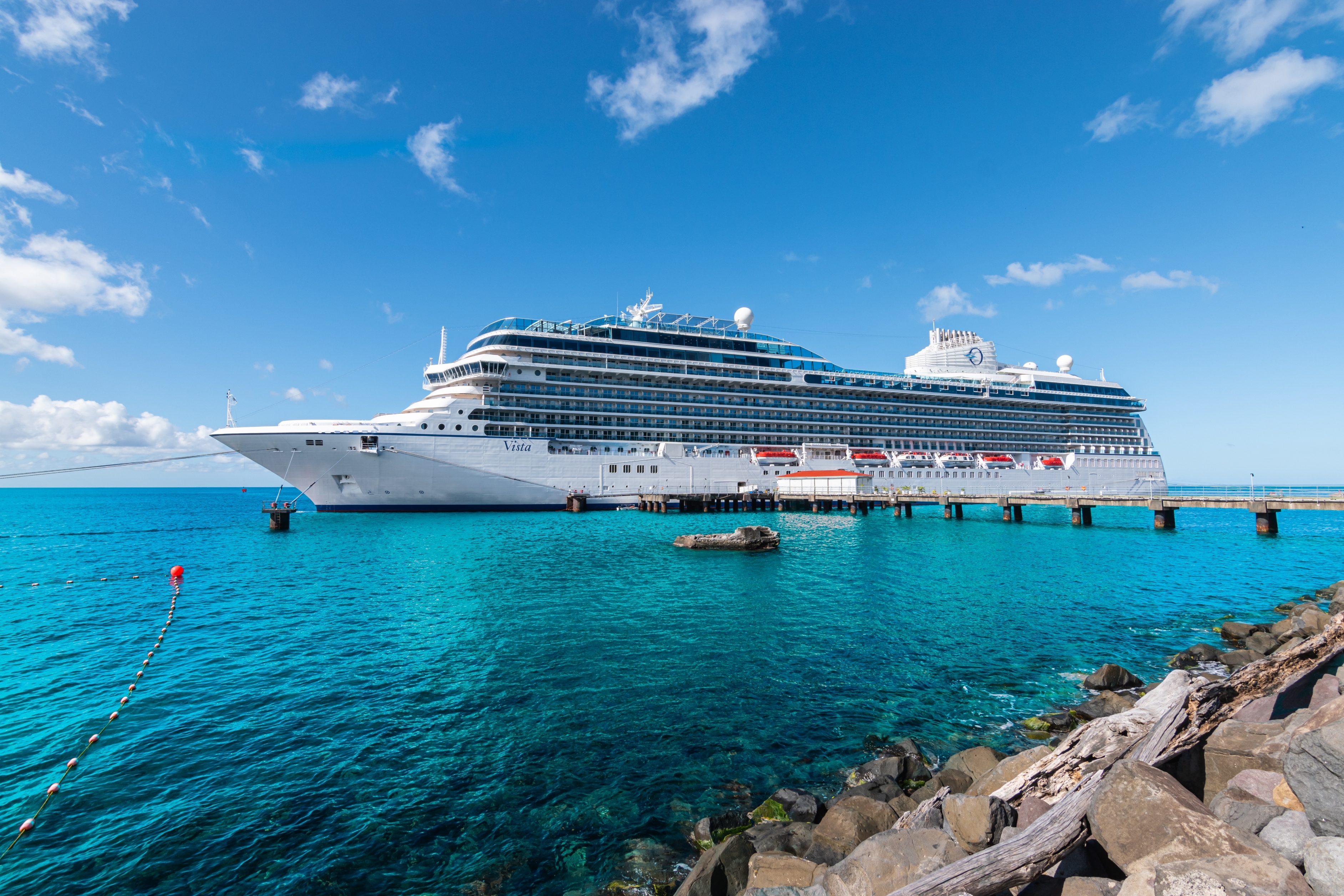 A large cruise ship docked at a pier in clear water under a bright sky, with rocks visible in the foreground