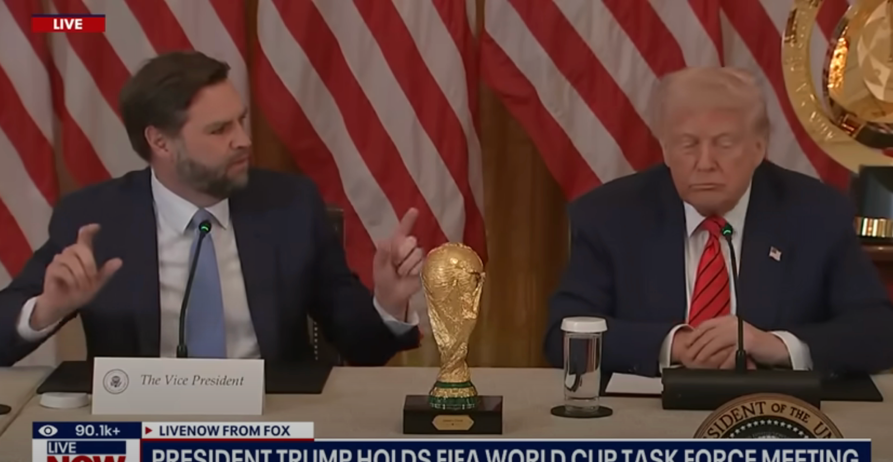 Two men seated at a table during a FIFA World Cup task force meeting. A trophy is placed between them