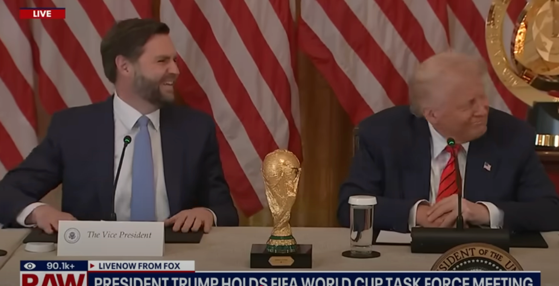 Two men seated at a table with a FIFA World Cup trophy, surrounded by U.S. flags at a meeting labeled "President Trump holds FIFA World Cup task force meeting."