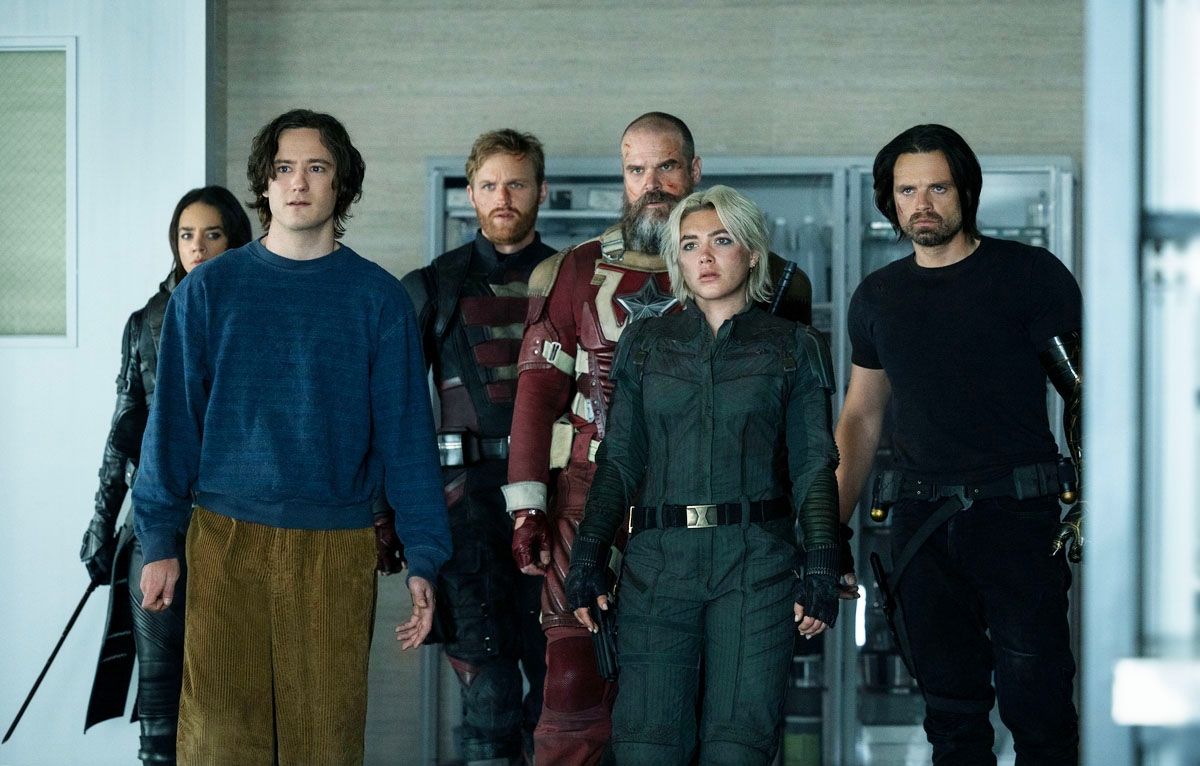 A group of actors in action-themed attire stands together, looking serious. They appear to be characters from a TV show or movie